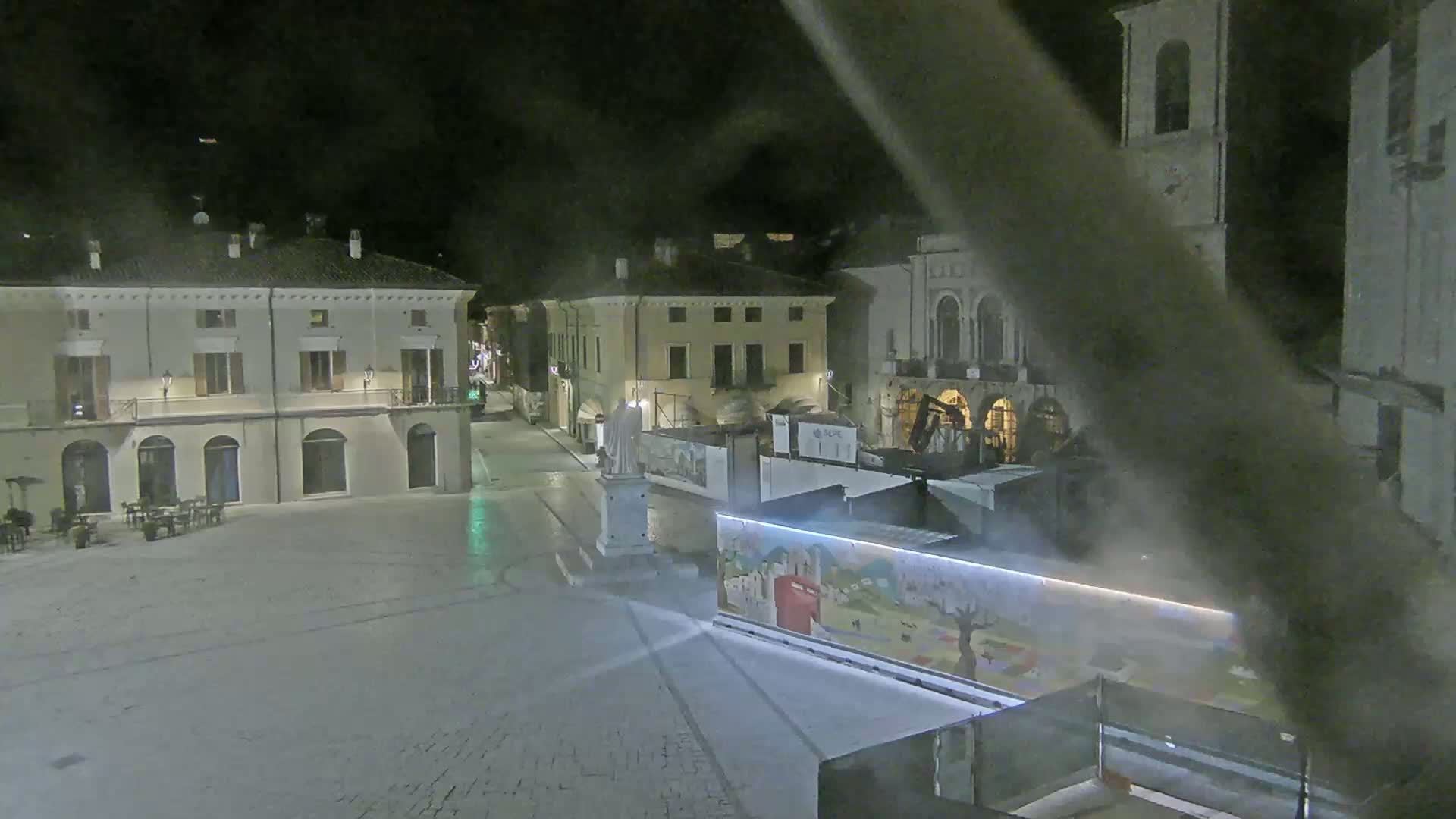 A nighttime view of a town square with several buildings, a statue, and construction barriers, under slightly hazy conditions.
