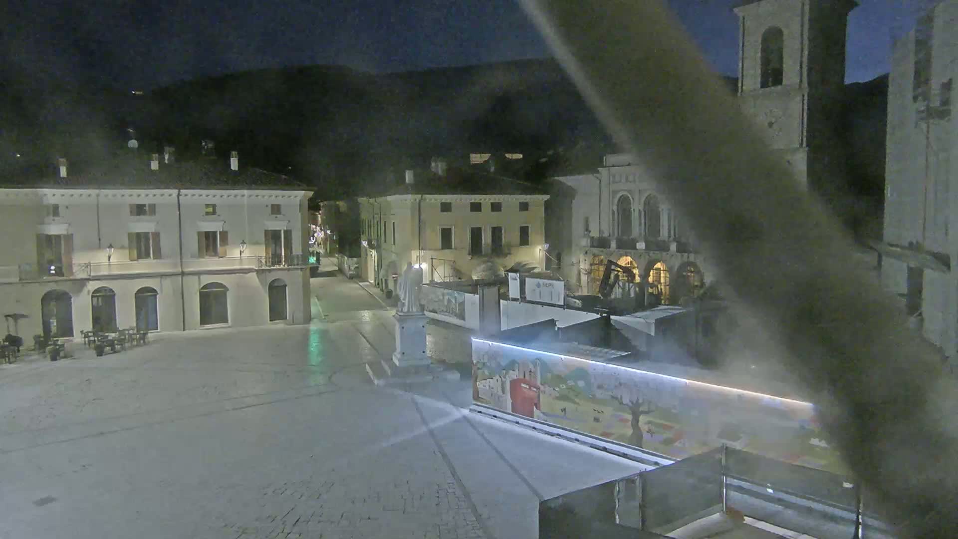 A nighttime view of a town square with several buildings, a statue, and a colorful mural on a temporary structure.