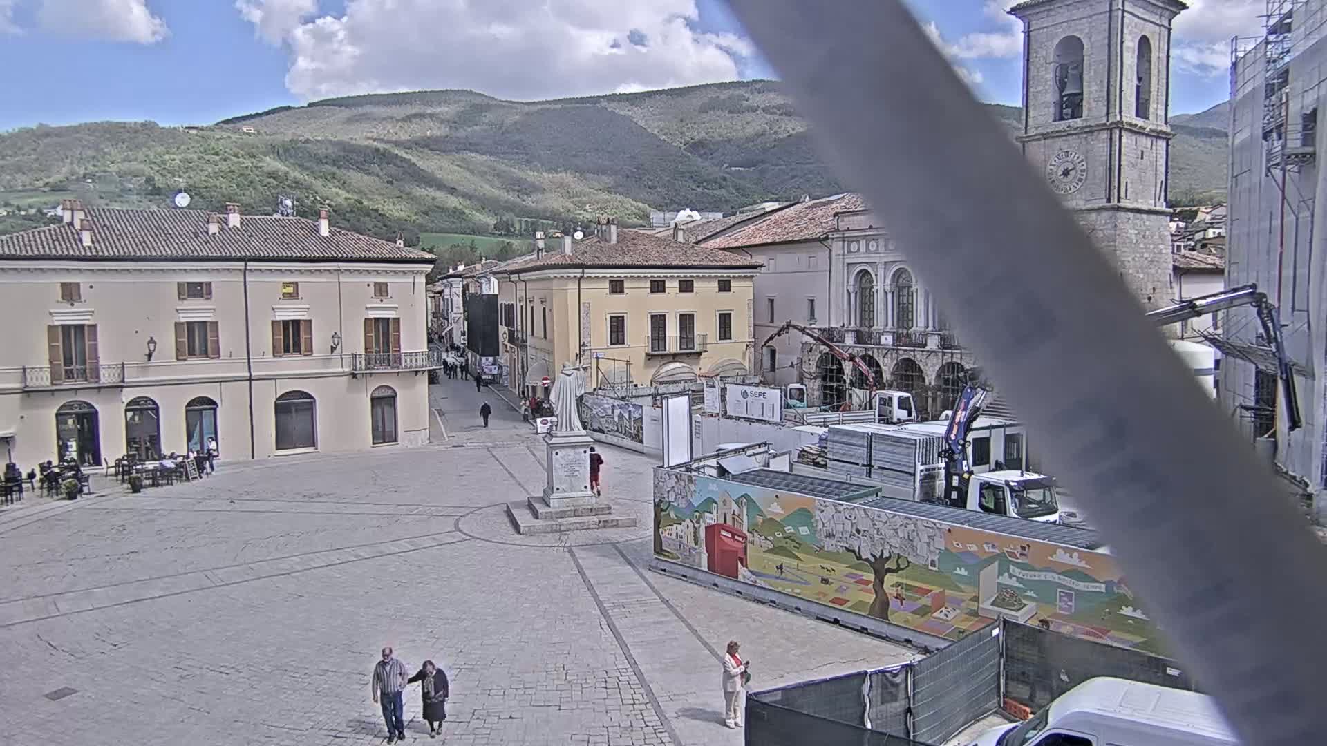 A mostly empty town square with several buildings, a statue, construction equipment, and a few pedestrians is viewed on a partly cloudy day in front of a mountain range.