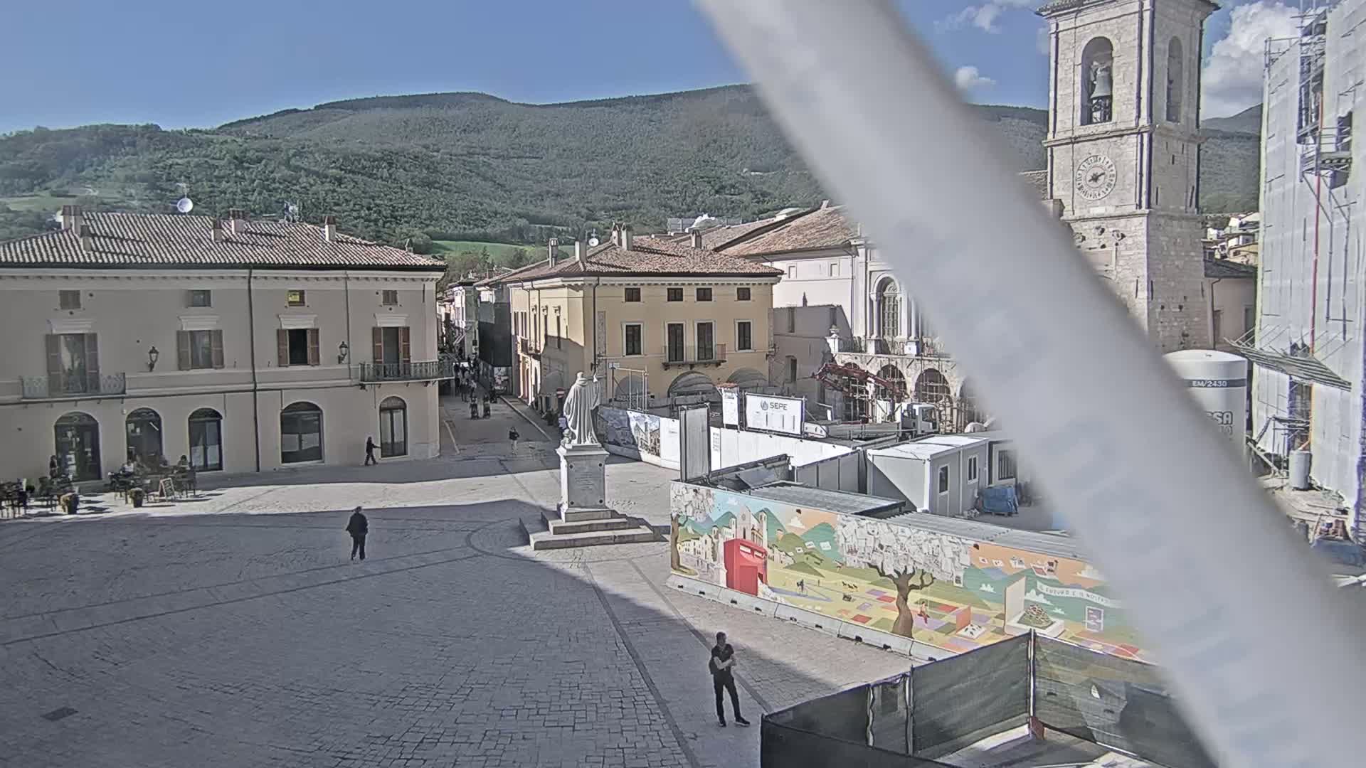 A mostly empty town square with several buildings, a statue, and construction underway, all under a clear, sunny sky with mountains in the background.