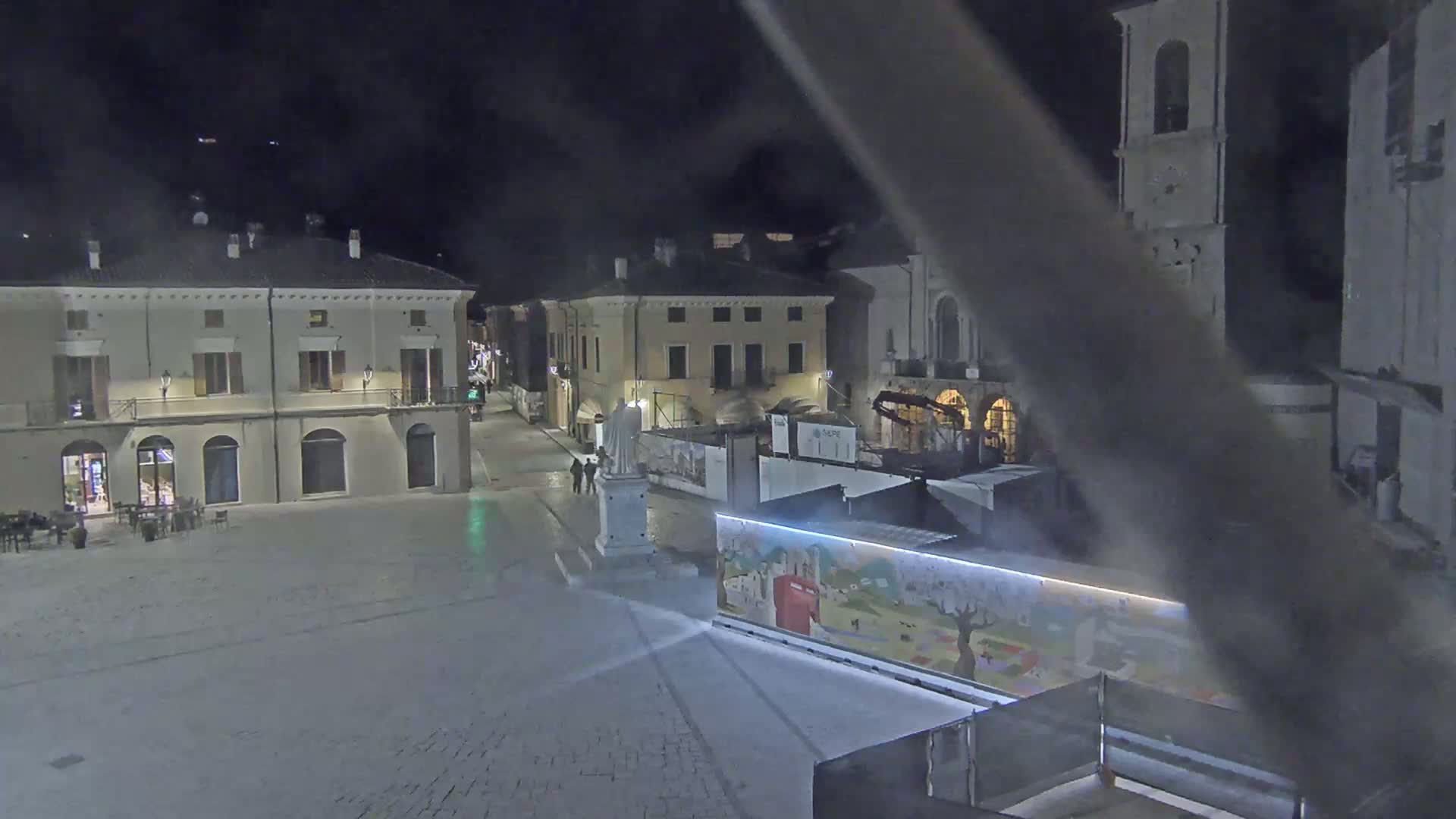 A mostly empty town square at night is seen under cloudy skies, with buildings lining the square and a statue in the center.