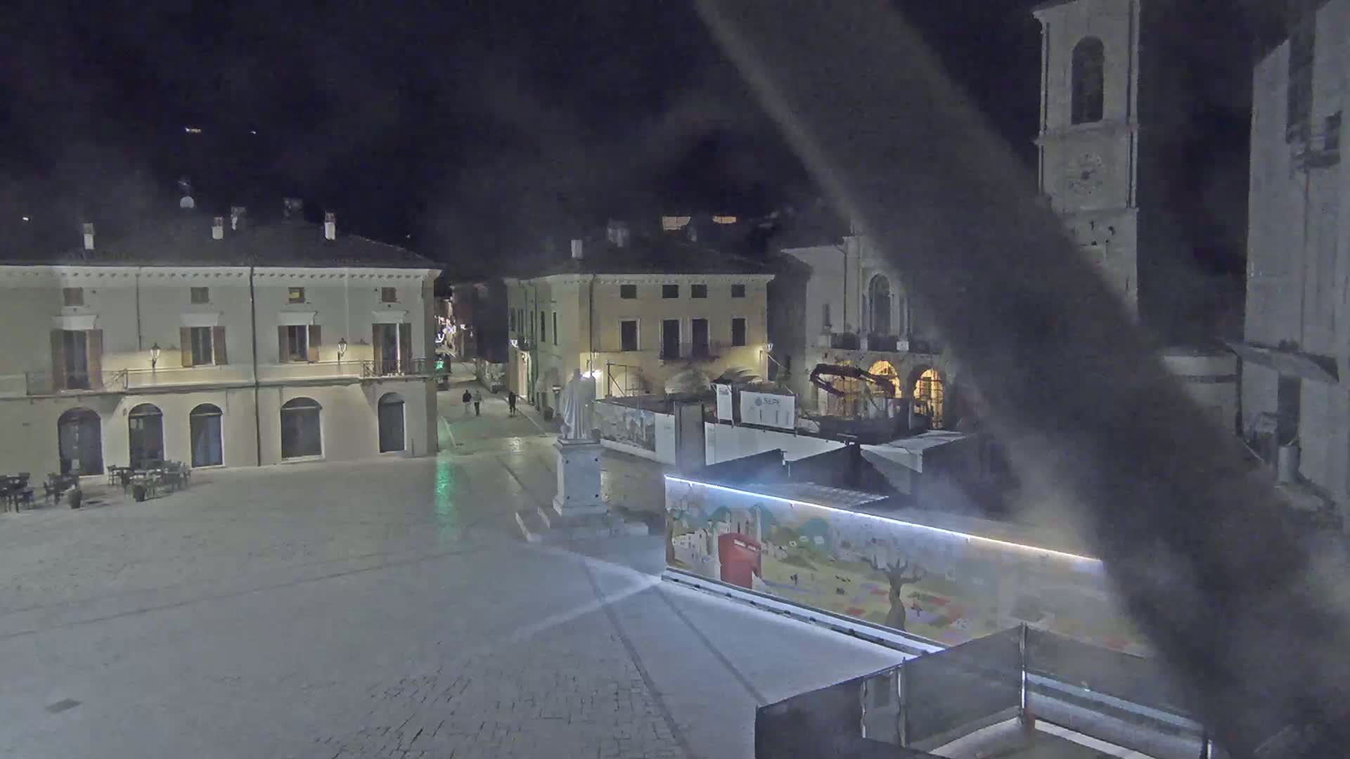 A nighttime view of an empty town square with several buildings, a statue, and construction barriers.