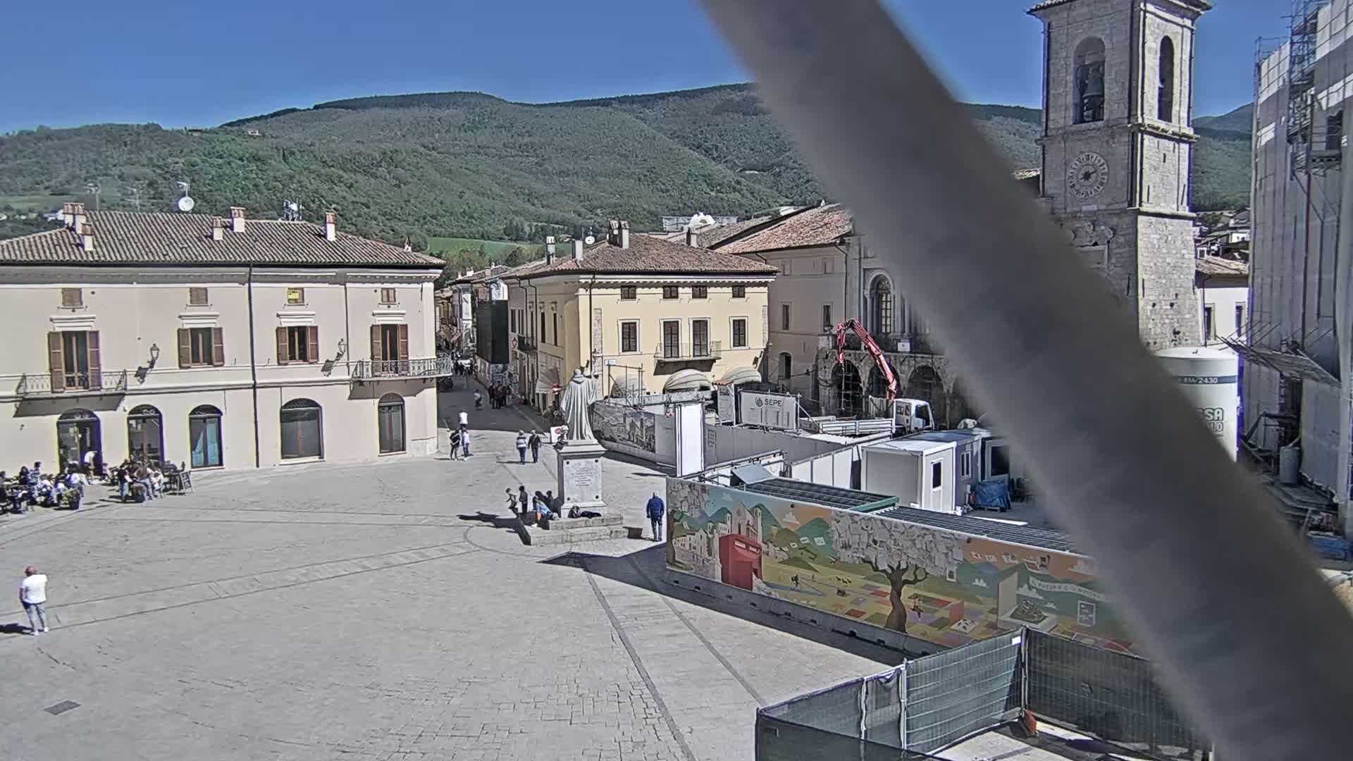 A town square with several buildings, a statue, and construction equipment, under a sunny sky with mountains in the background.
