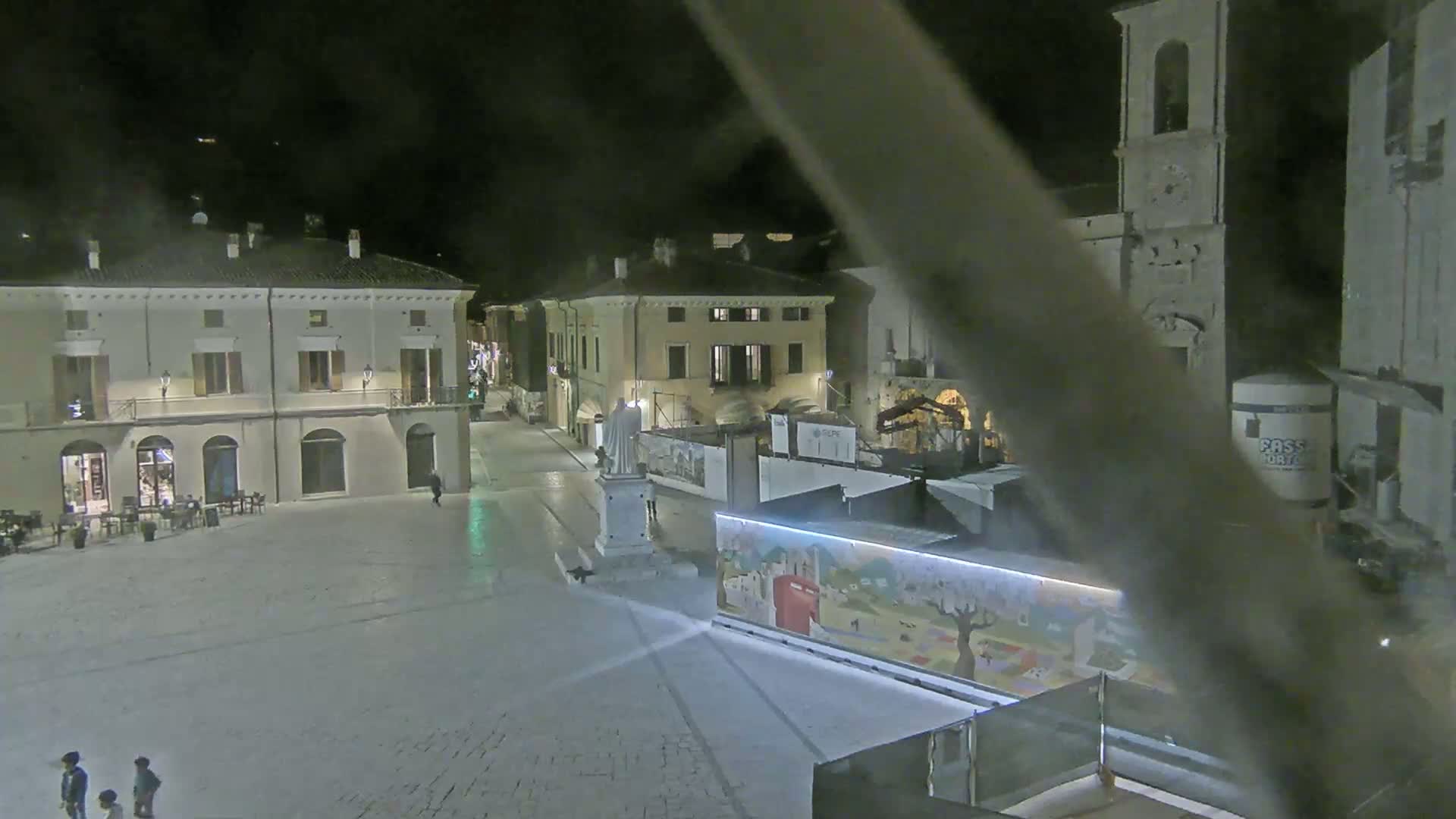 A nighttime view of a town square with several buildings, a statue, and construction barriers, under clear skies.