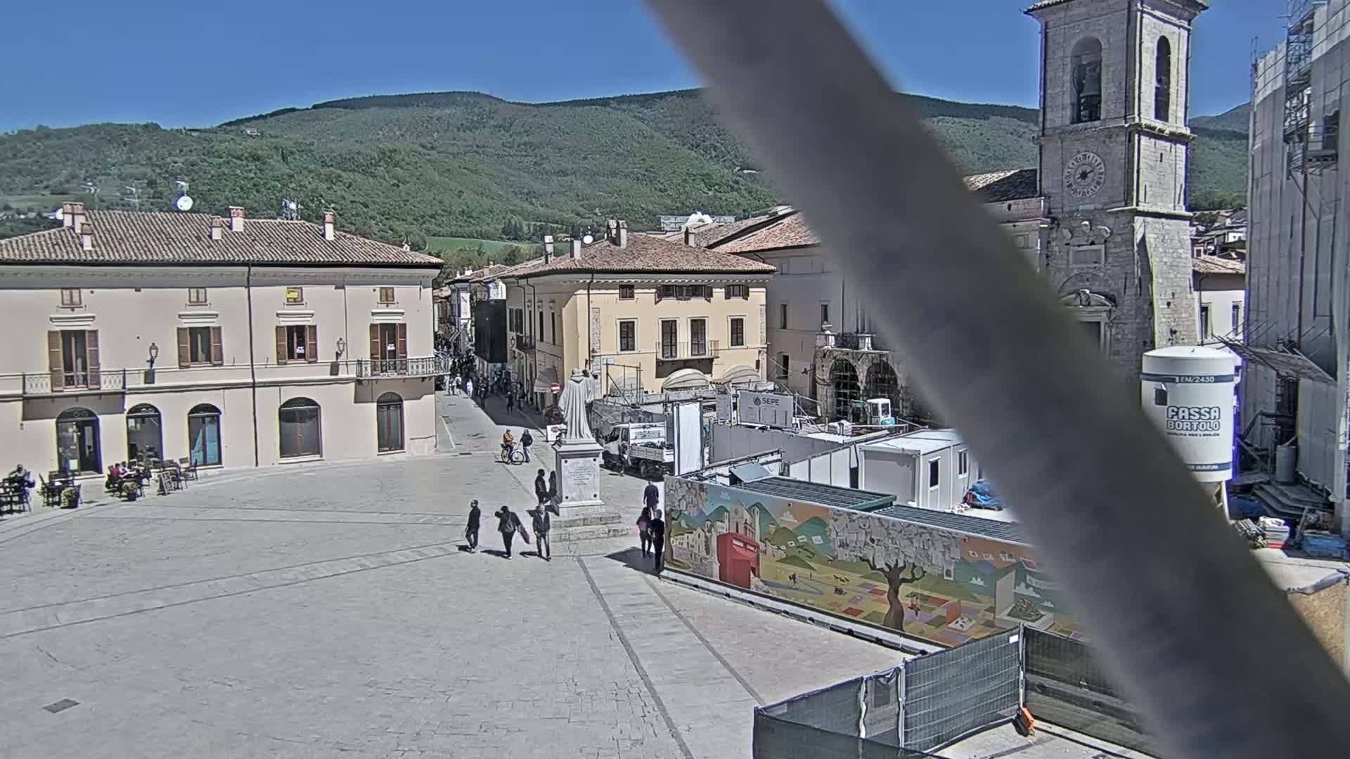 A sunny town square with several buildings, a statue, and a few people walking around, partially obscured by a metal pole, with mountains in the background.