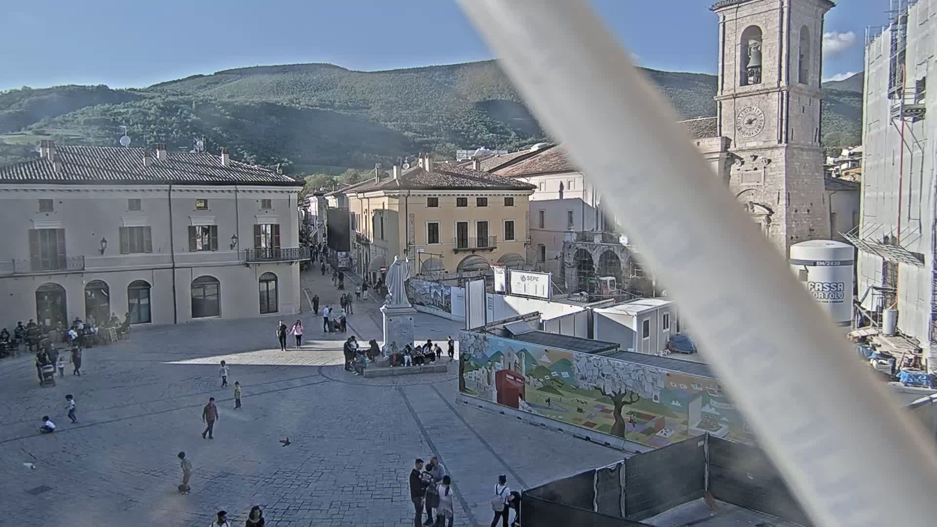A town square, under sunny skies and surrounded by historic buildings, is populated by several people, some of whom are sitting, while construction is underway in the background near a tall bell tower.