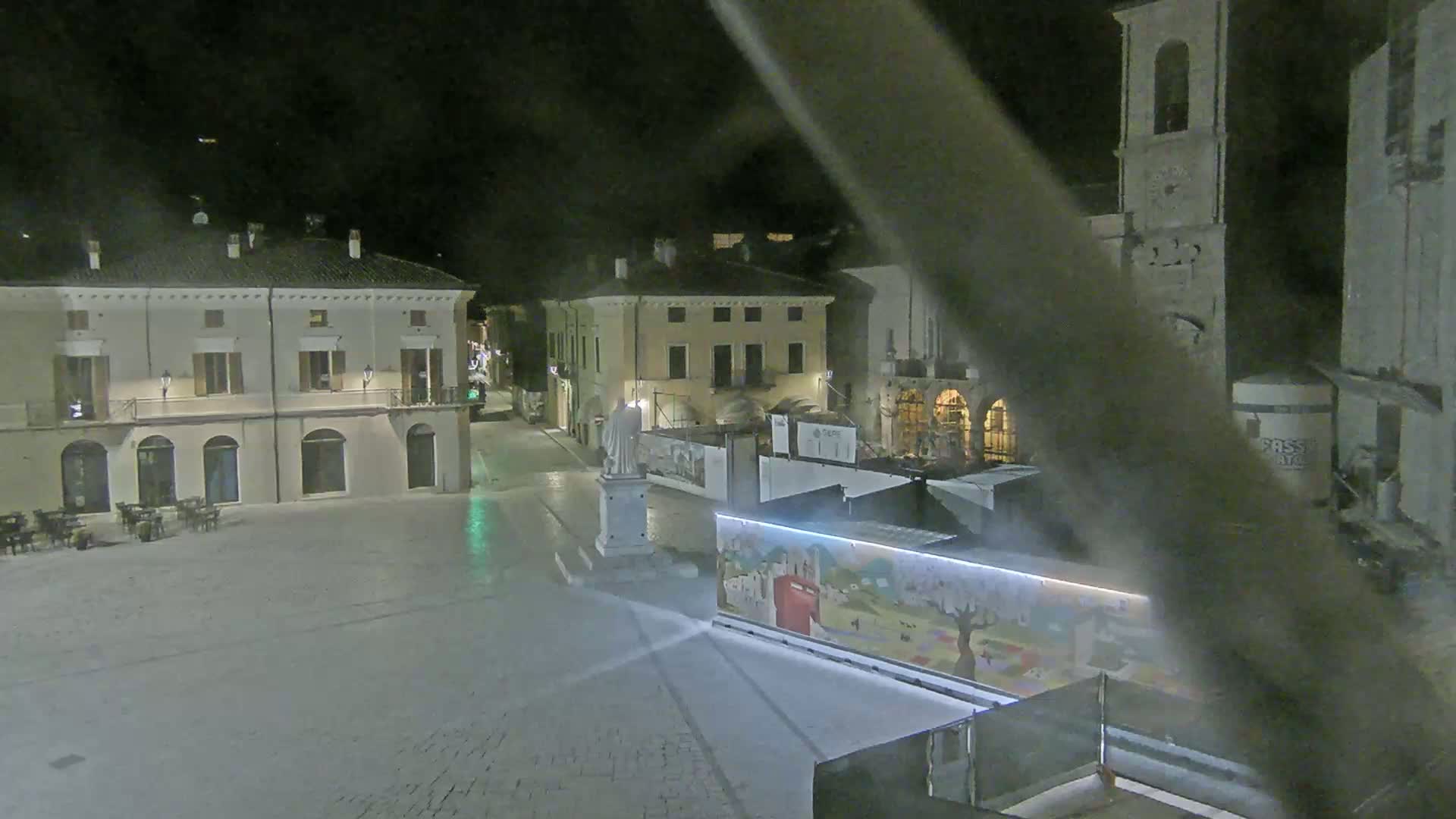 An empty town square at night, illuminated by streetlights, features several buildings, a statue, and a painted structure.