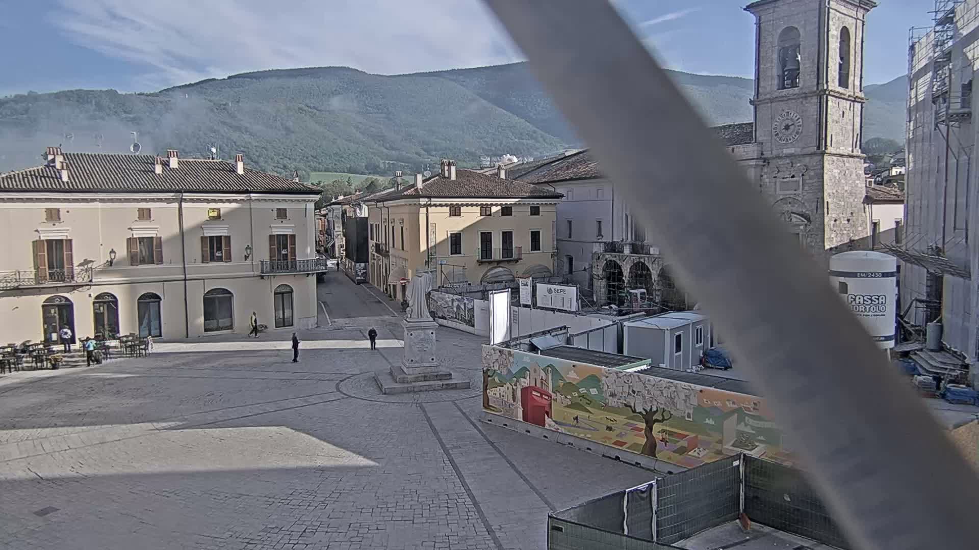 A mostly empty town square, under a partly cloudy sky, is partially obstructed by construction and features buildings, a statue, and mountains in the background.