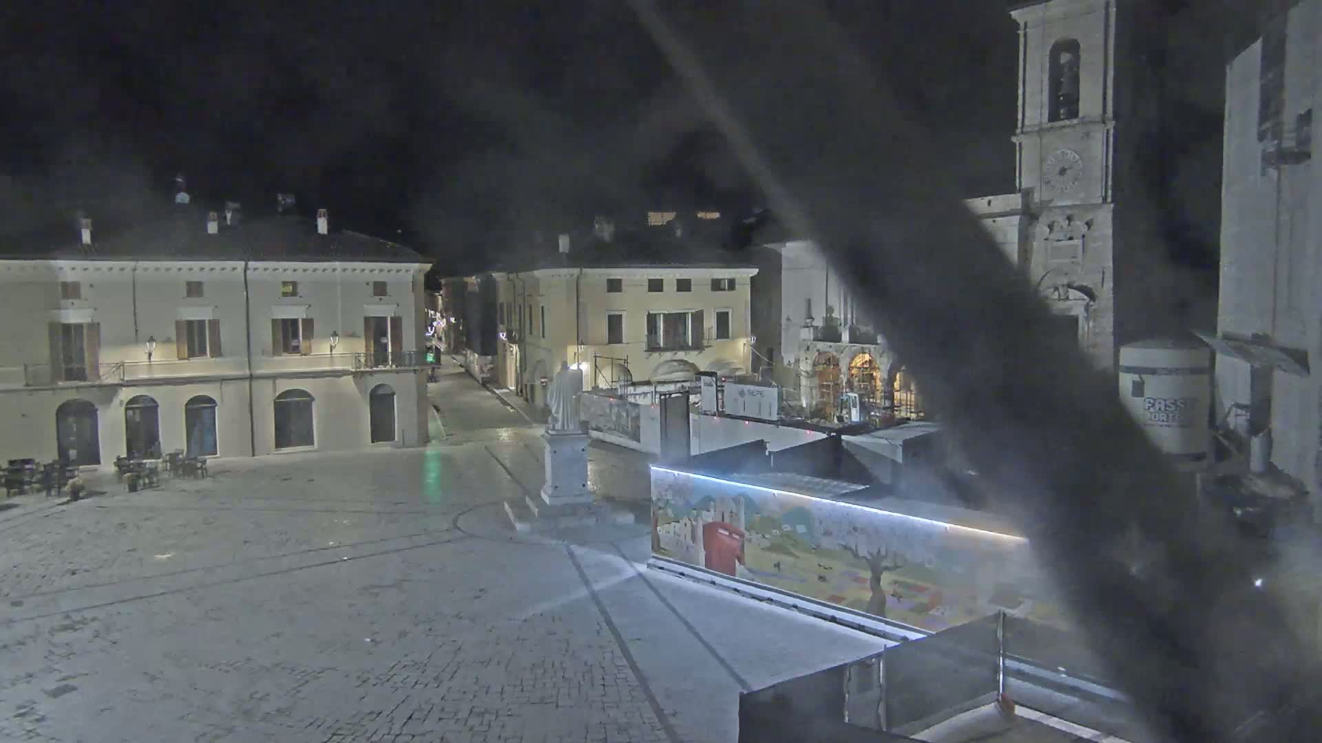 A nighttime view of an empty town square with several buildings surrounding it, including a tall bell tower, under dark, possibly overcast conditions.