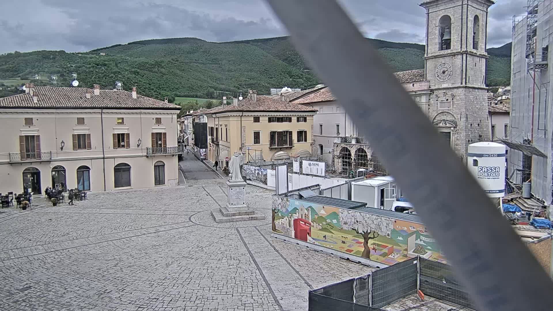 A stone town square under an overcast sky, featuring several buildings, a statue, and construction barriers.