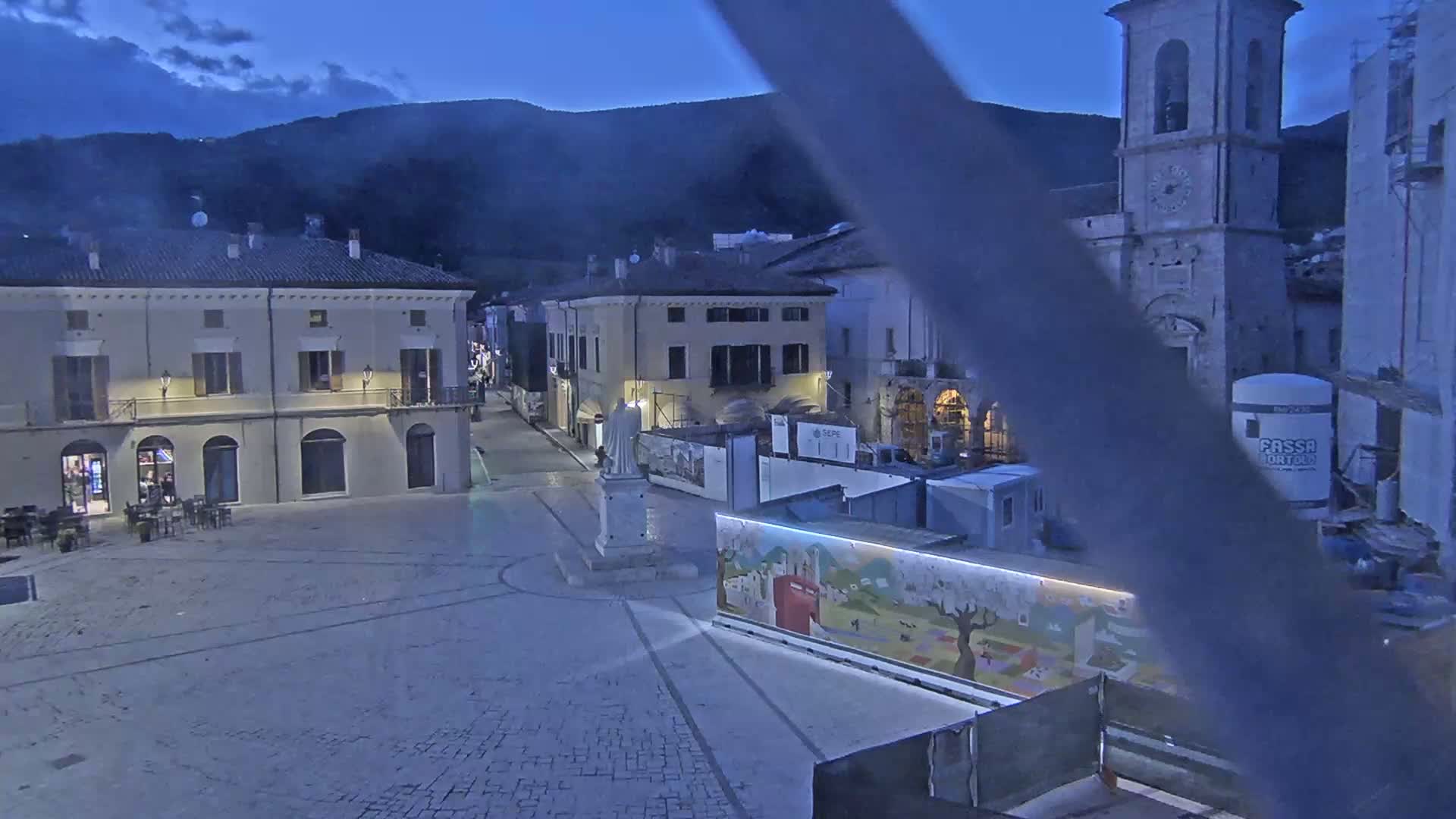 A twilight view of a town square, featuring several buildings, a statue, construction, and a painted wall, under a partly cloudy sky.