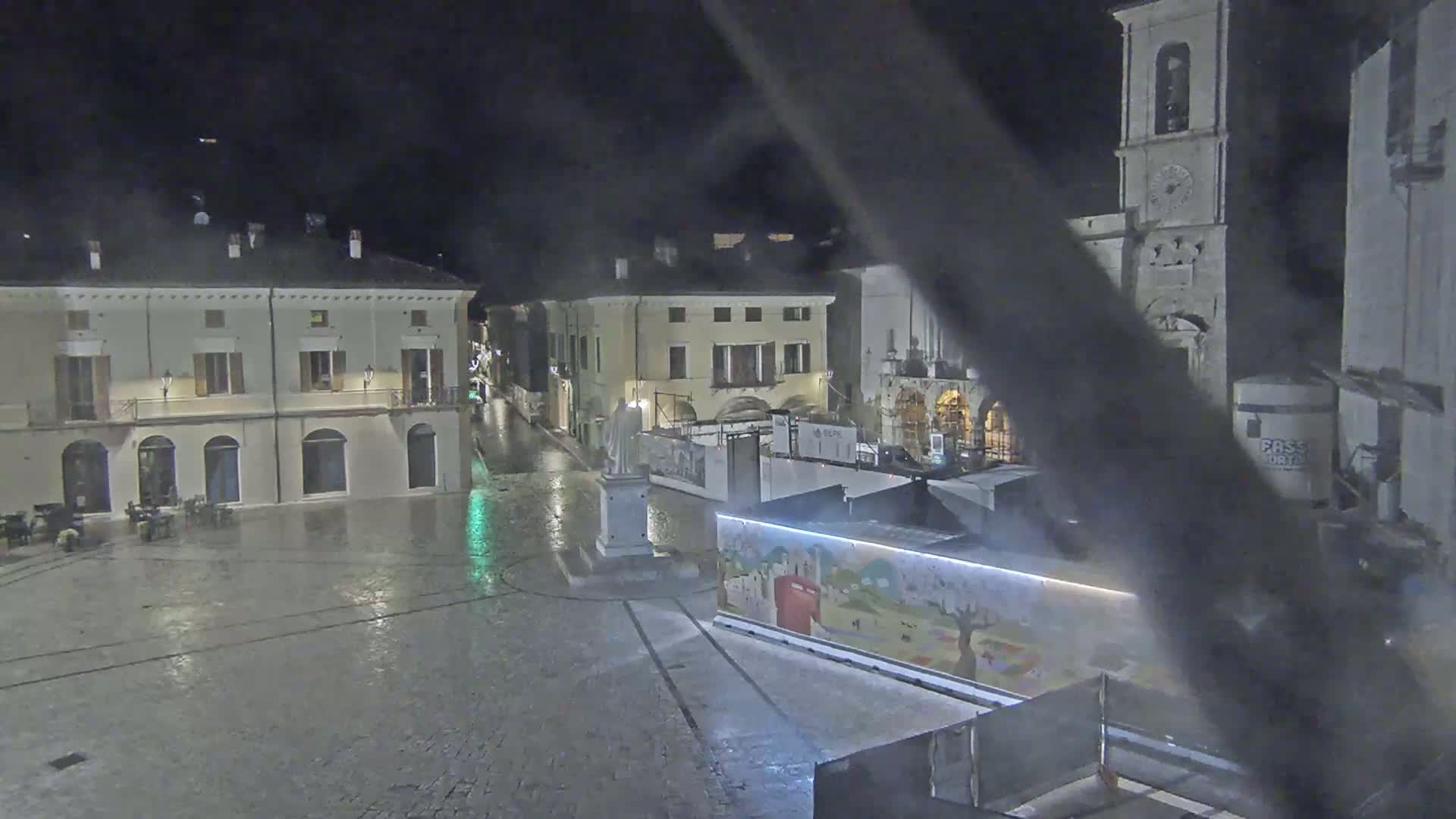 A rain-slicked town square at night, surrounded by old buildings, with a statue in the center and construction barriers partially obscuring a painted wall.