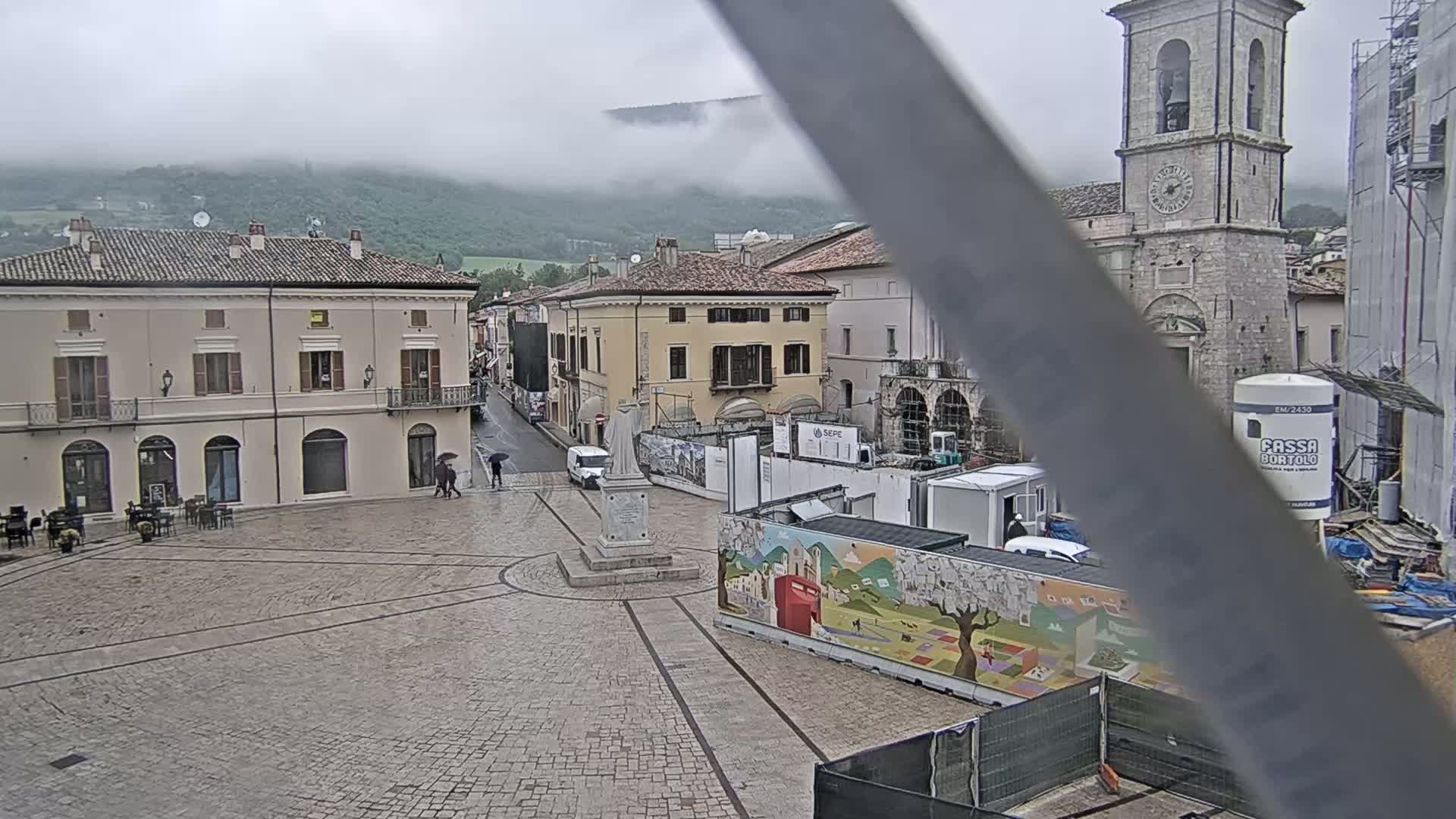 A rain-soaked town square, surrounded by buildings and a partially obscured church tower, is nestled beneath a foggy mountain.