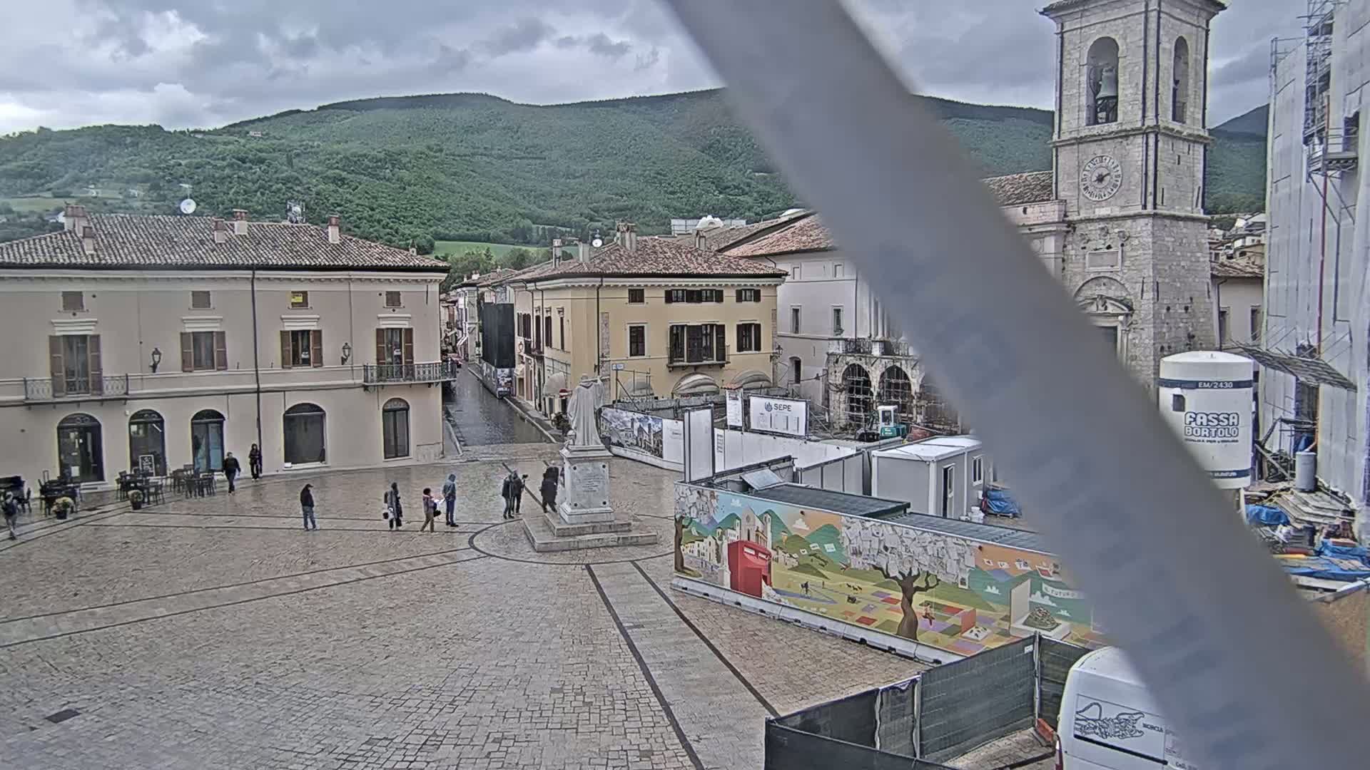 A town square with several buildings, a statue, and some people, under an overcast sky, with construction taking place.