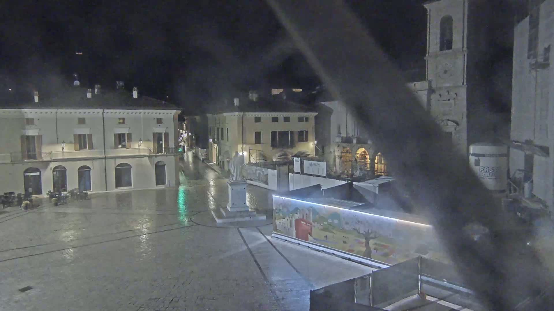 A rain-slicked town square at night, surrounded by buildings, features a statue on a pedestal and construction barriers with artwork.
