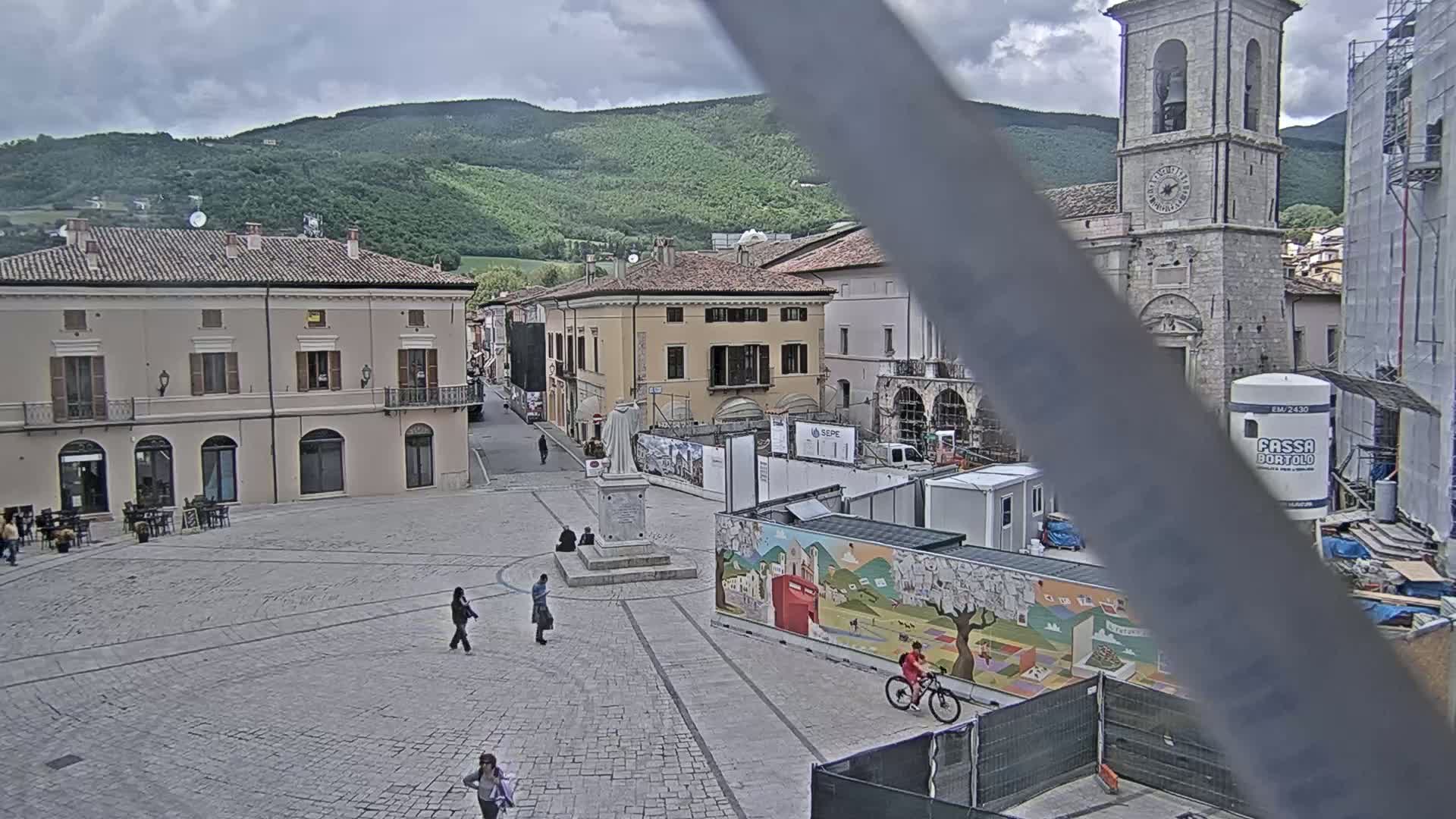 A town square, under a cloudy sky, is partially under construction with scaffolding and barriers, featuring a stone church tower in the background and several people walking around.