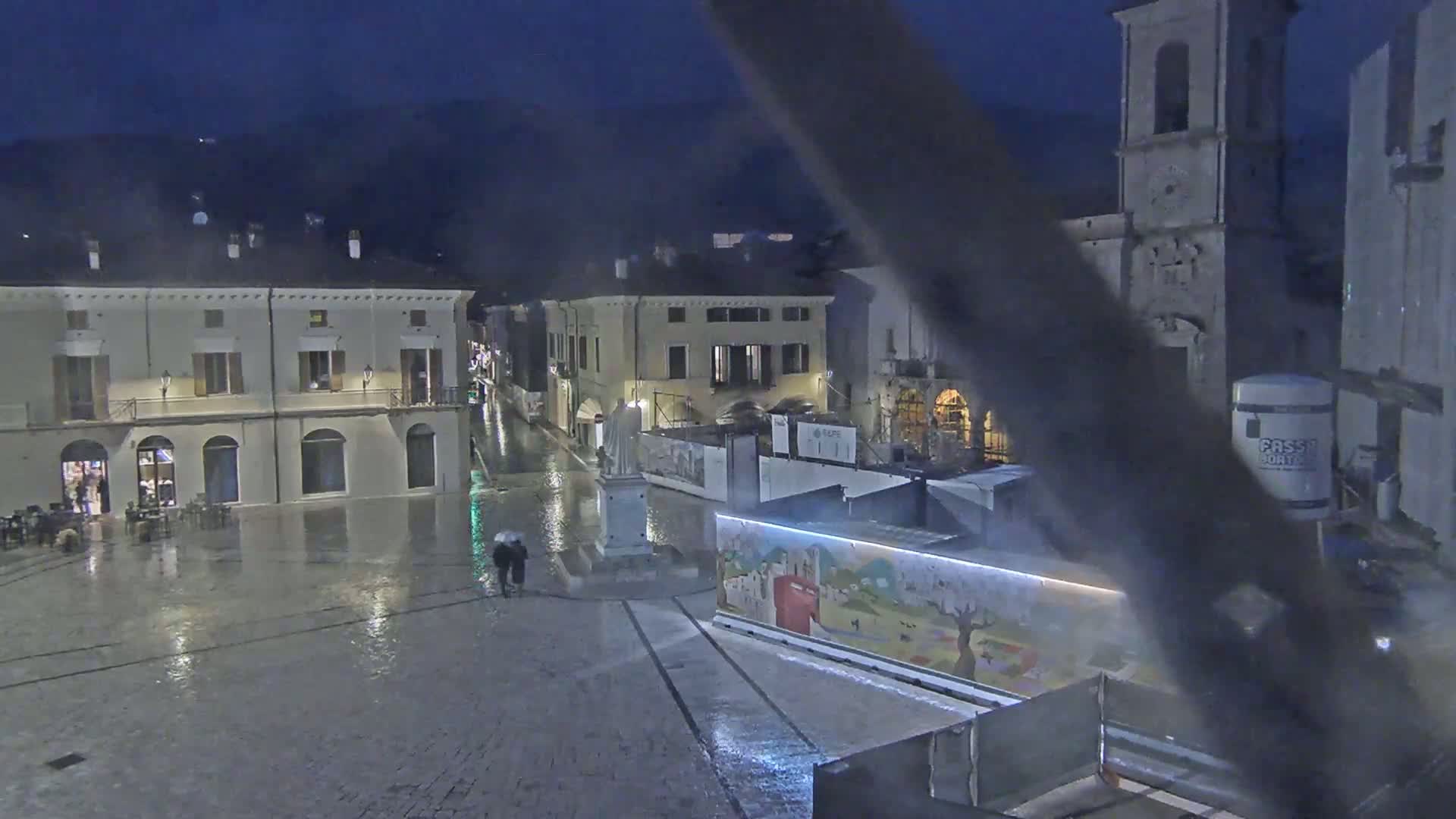 A rain-soaked town square at night, featuring several buildings, a statue, and two people under an umbrella.