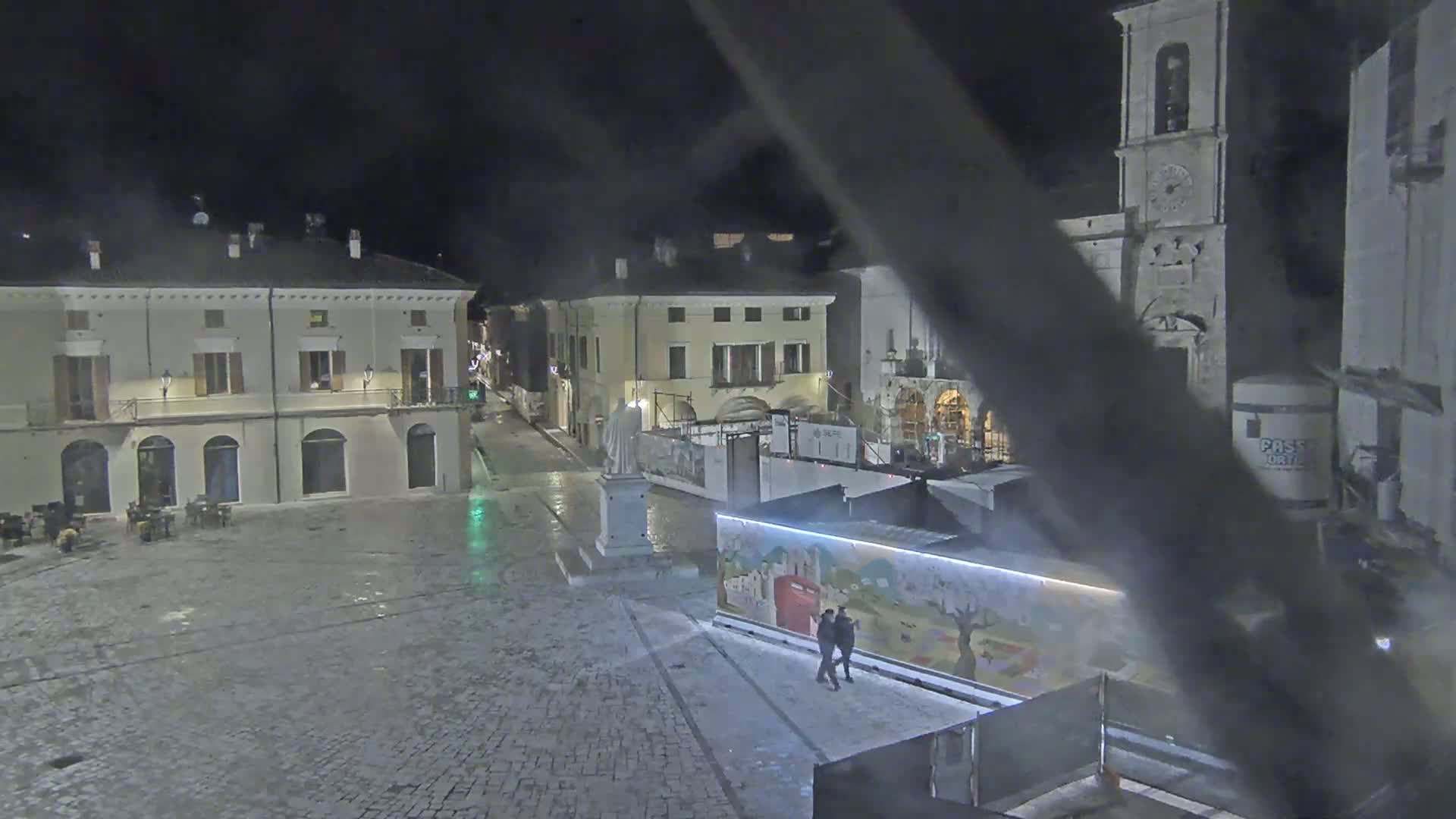 A nighttime view of a town square, showing several buildings, a statue, and a few people walking near a brightly colored wall; the ground is wet.