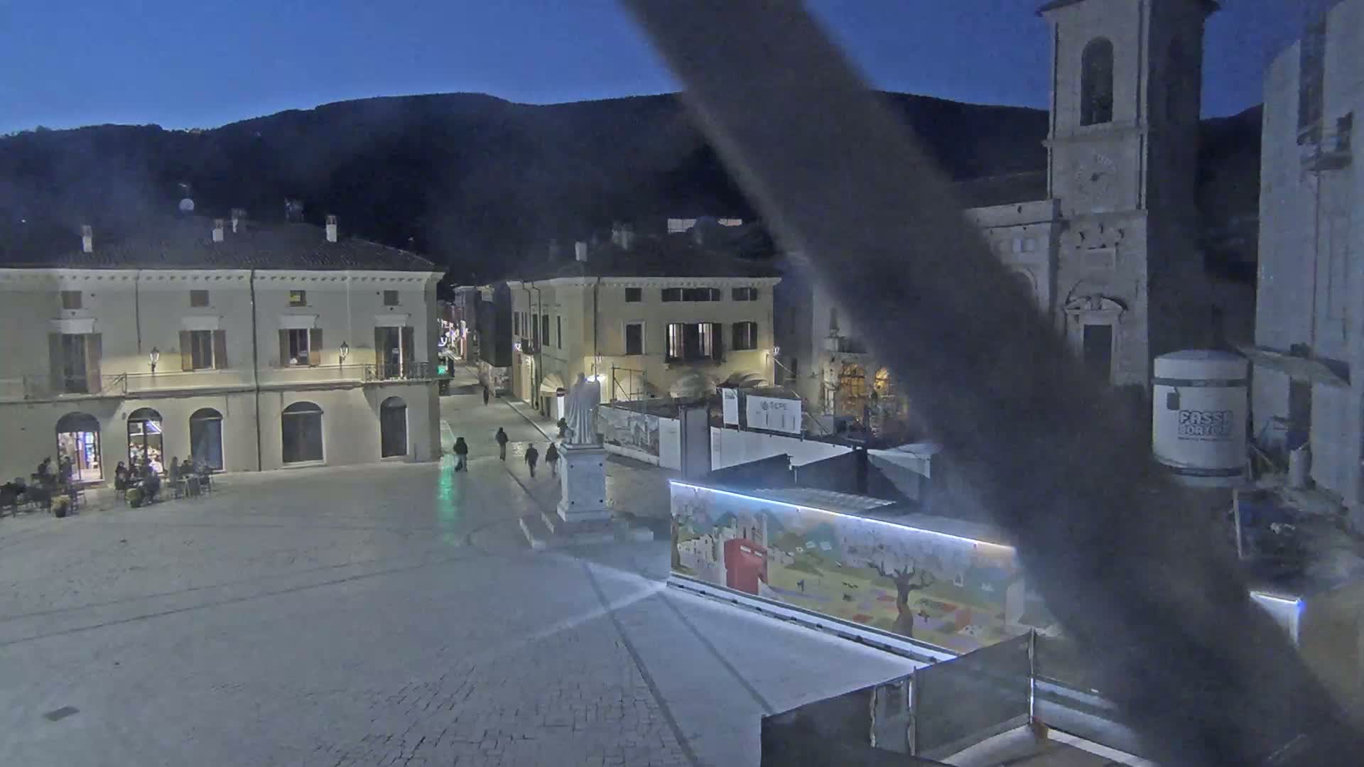 A mostly empty town square at night, under a dark blue sky, is partially obstructed by construction and features several buildings, including a church.