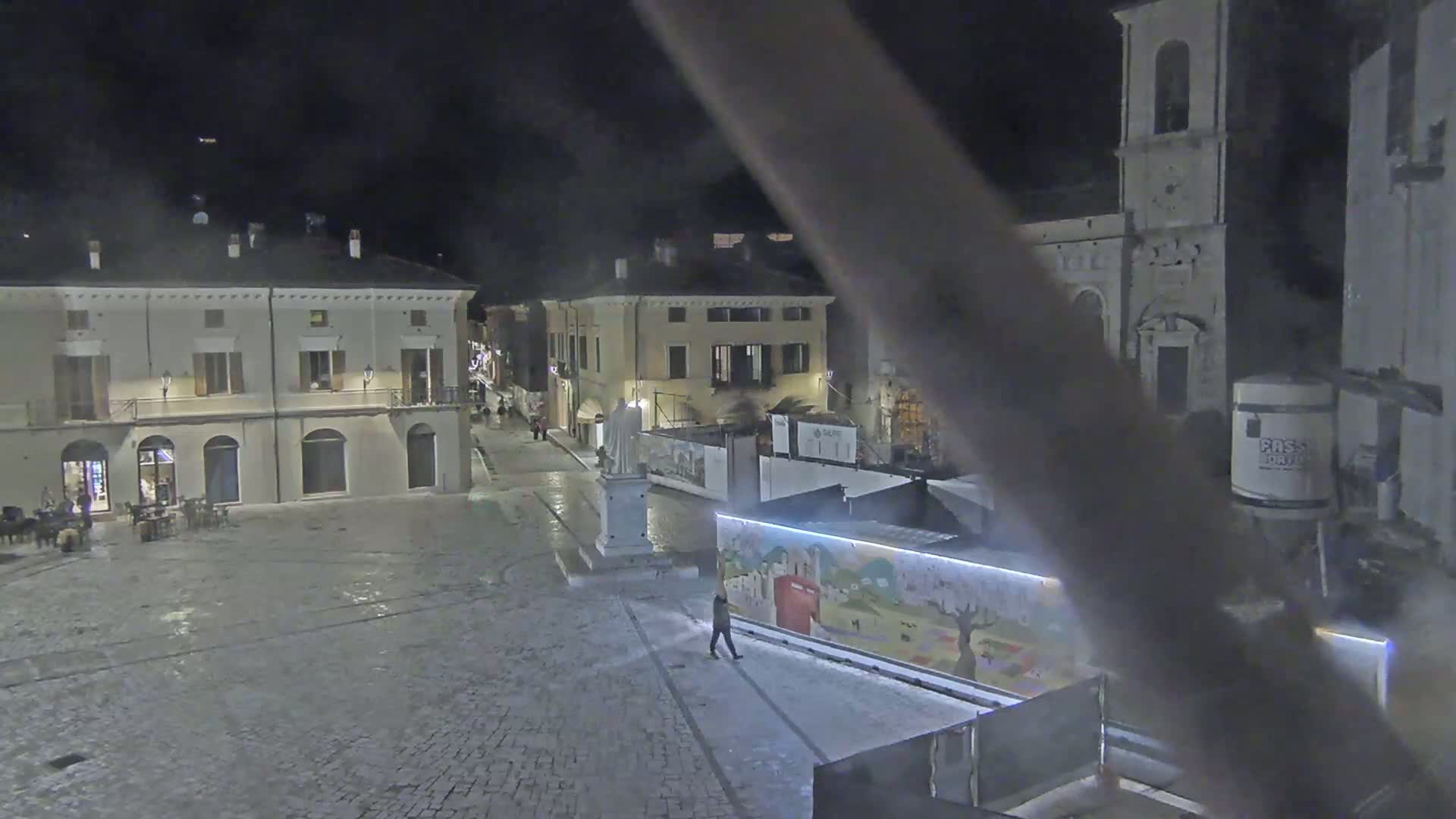 A nighttime view of a cobblestone town square with several buildings, a statue, and construction barriers.