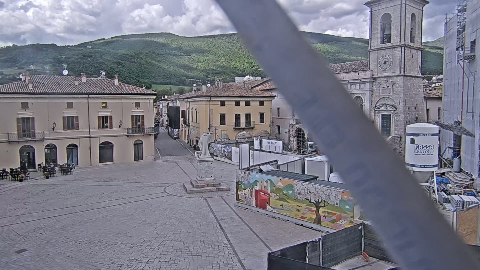 A mostly cloudy day reveals a town square with several buildings, a statue, and construction, all set against a backdrop of green hills.