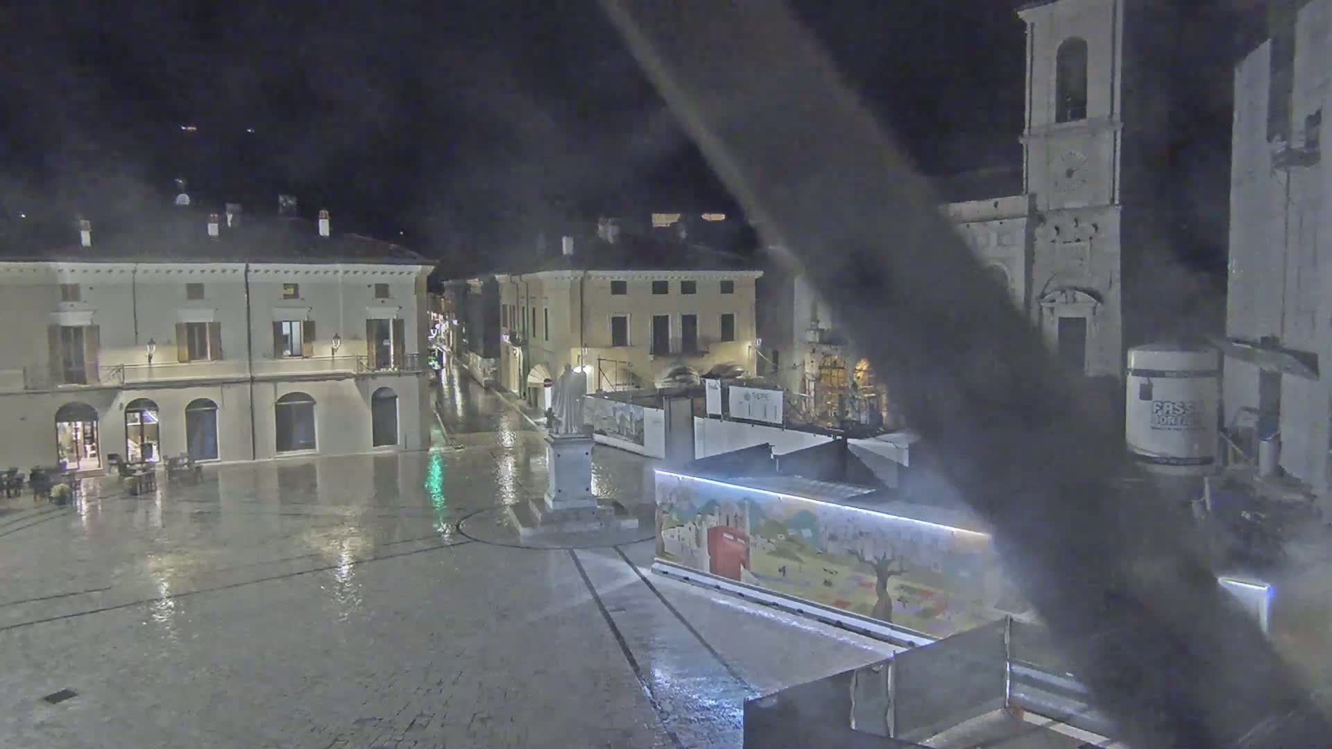 A rain-slicked town square at night, surrounded by historic buildings, one of which is a church with a tall bell tower, with construction or renovation work underway in the square.