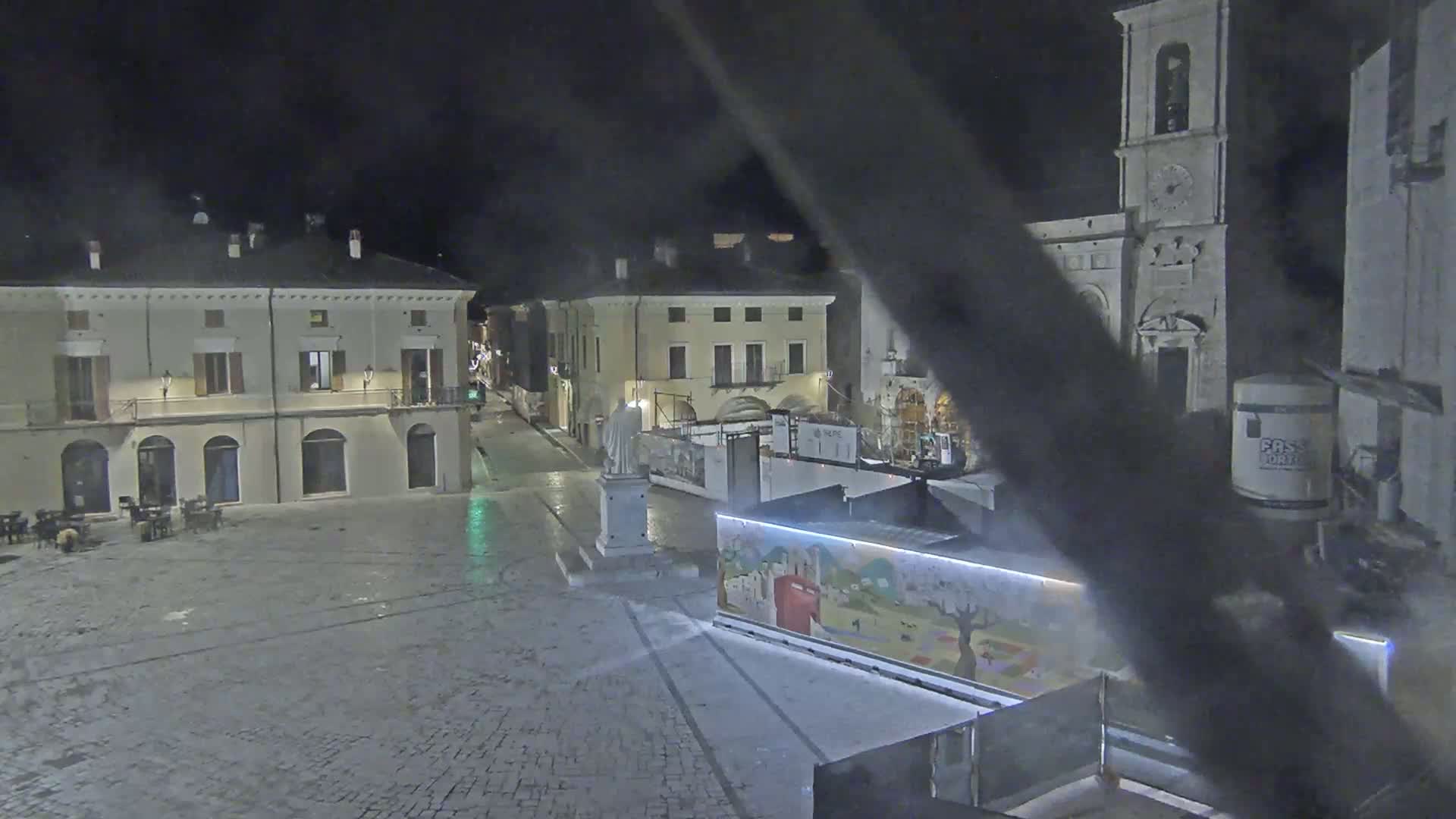 A nighttime view of a cobblestone town square with buildings, a statue, and construction barriers under dark, possibly overcast conditions.