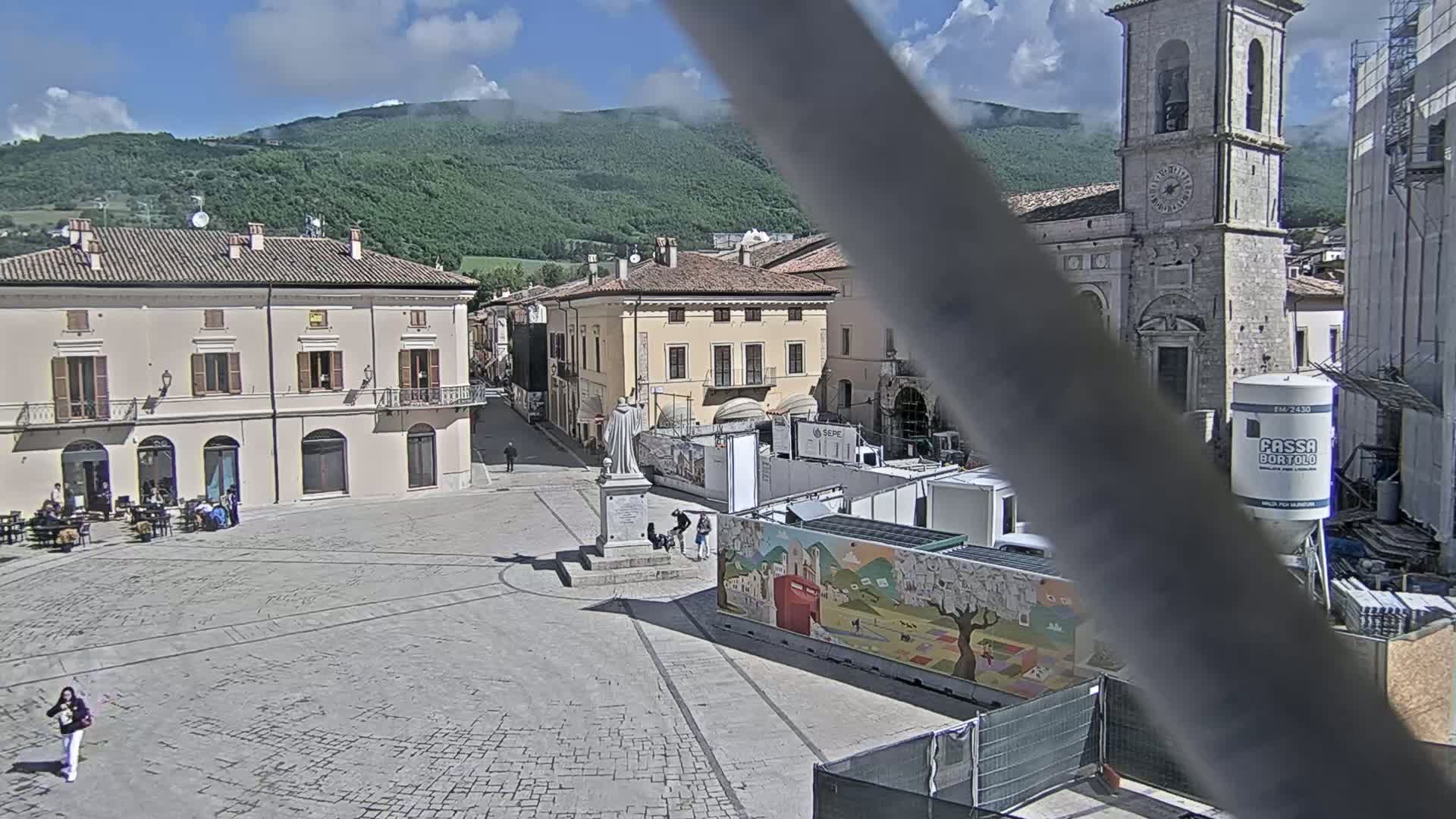 A mostly sunny town square with several buildings, a statue, and construction equipment is visible, with a mountain range in the background.