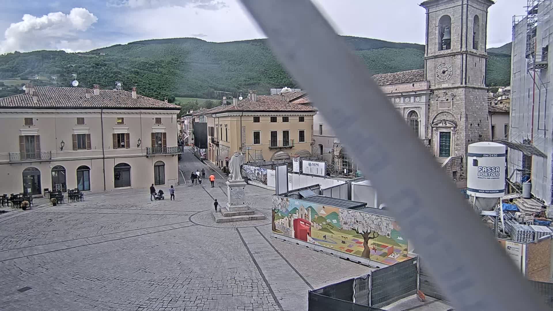 A mostly empty cobblestone town square is under a partly cloudy sky, with several buildings surrounding it and construction work visible near a tall church tower in the background.