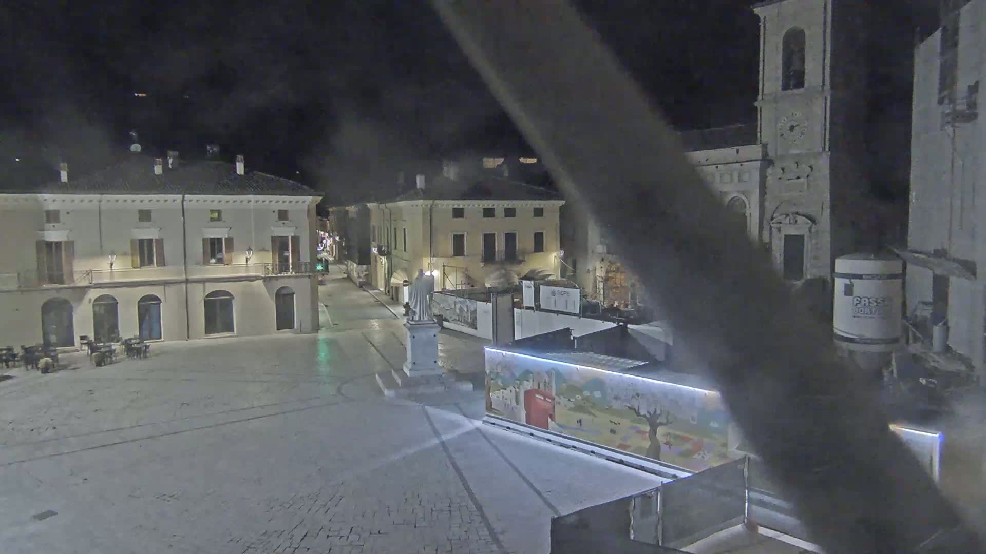 A nighttime view of a town square with several buildings, a statue, and construction barriers.