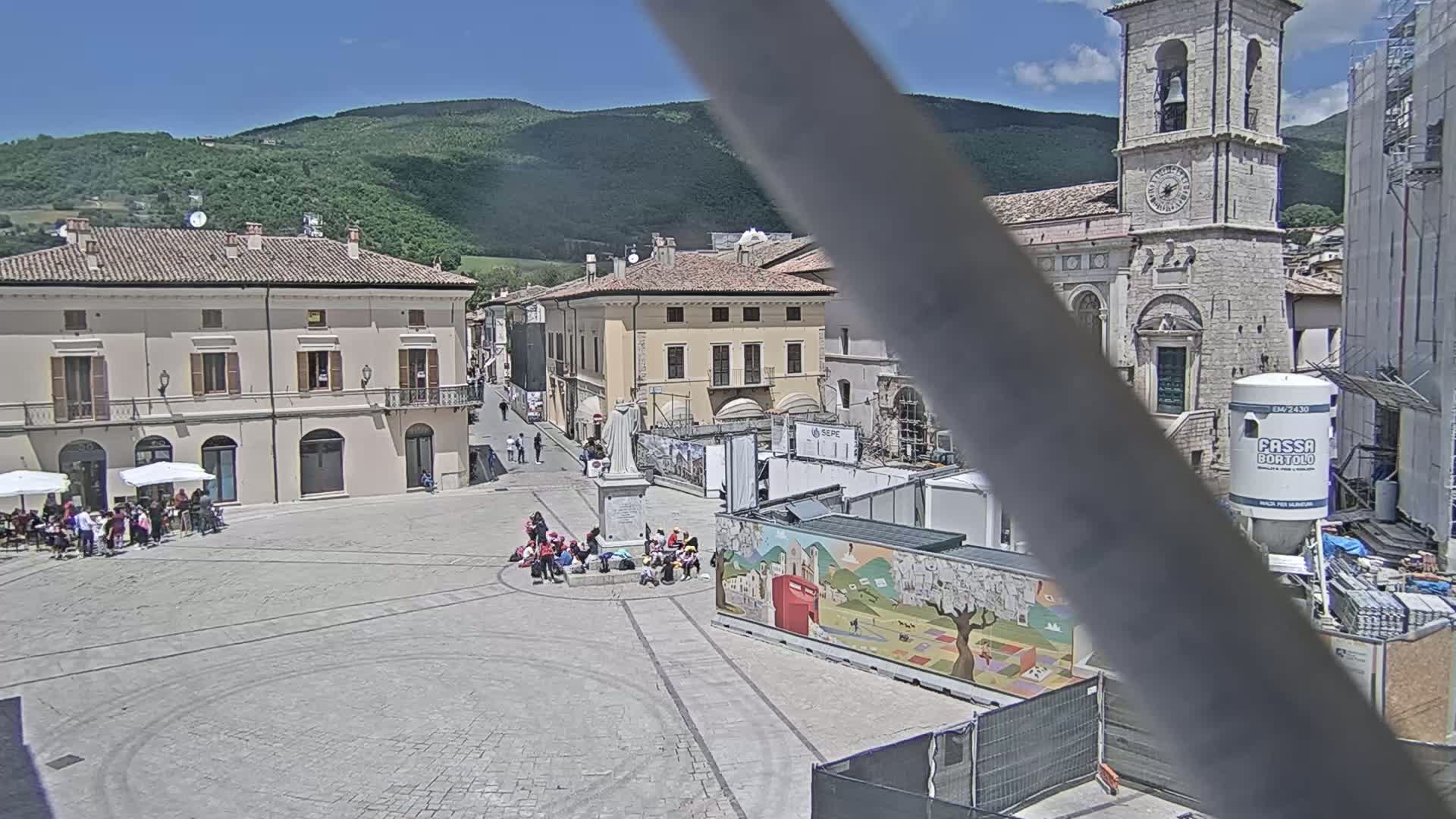 A town square with several buildings, a statue, and people sitting under umbrellas on a sunny day, with mountains in the background.