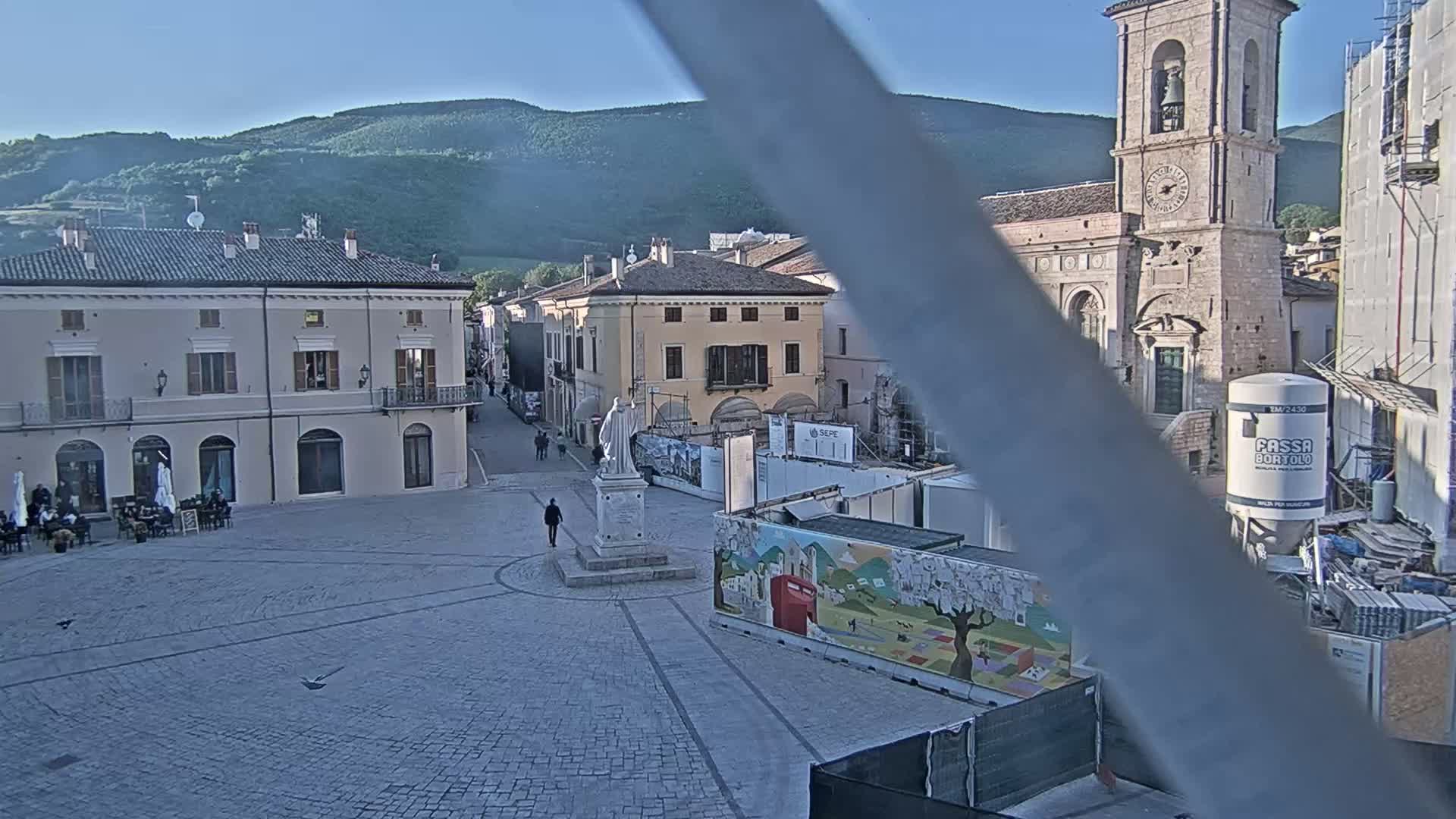 A town square with several buildings, a statue, and construction materials, under a clear blue sky, is visible against a backdrop of distant hills.