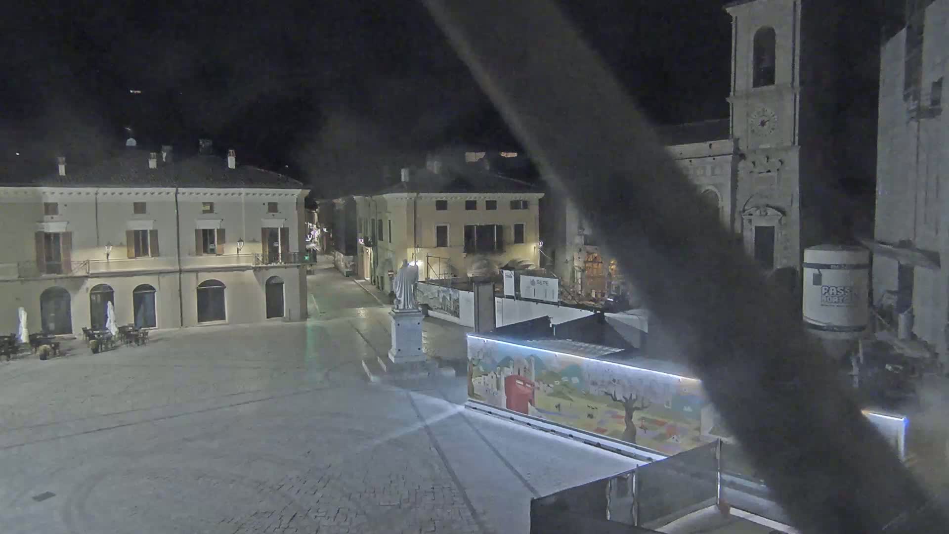 A nighttime view of a town square with several buildings, a statue, and construction barriers, under a clear, dark sky.