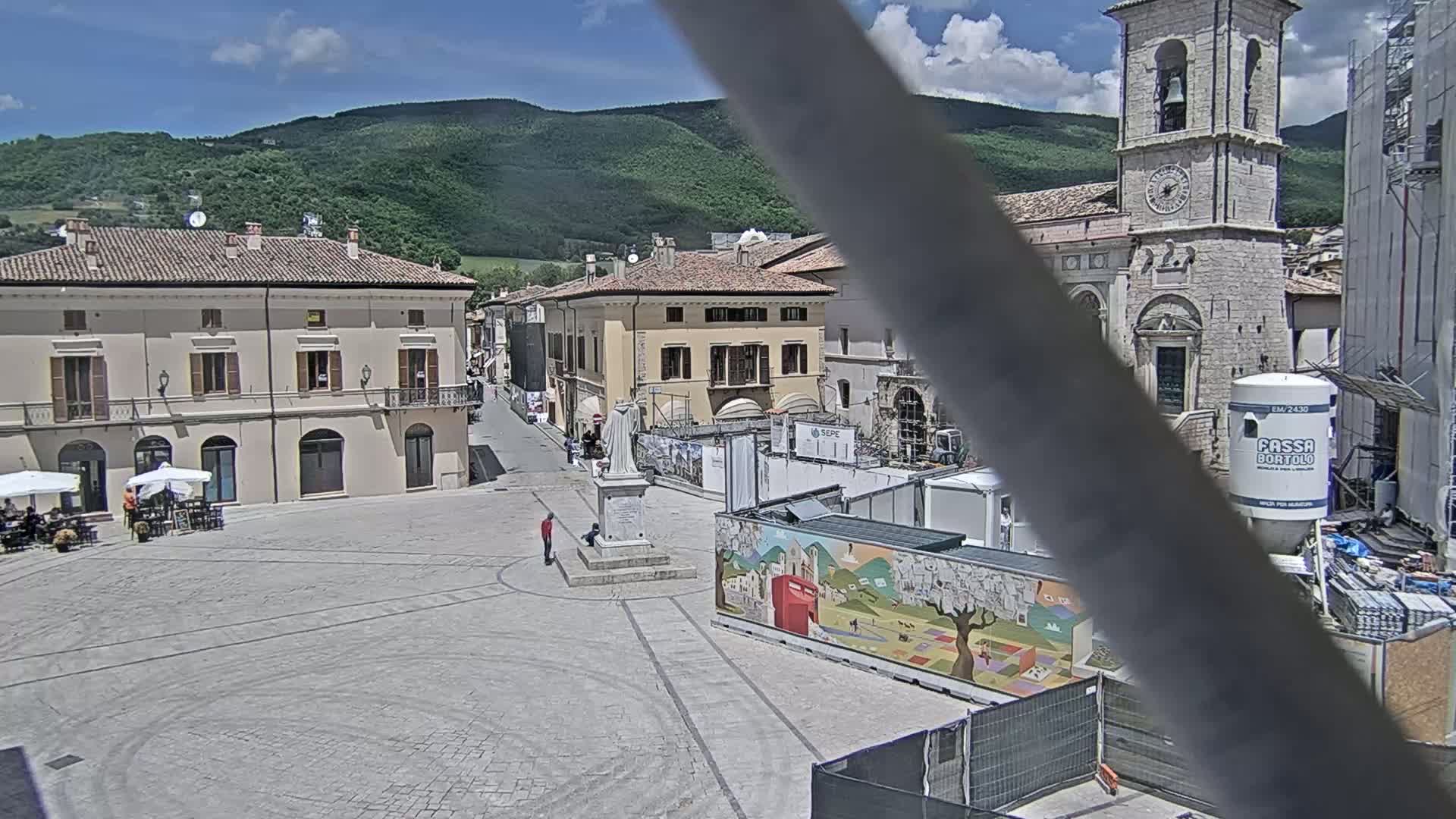 A town square in a sunny, mountainous region features several buildings, a statue, construction barriers, and some people.
