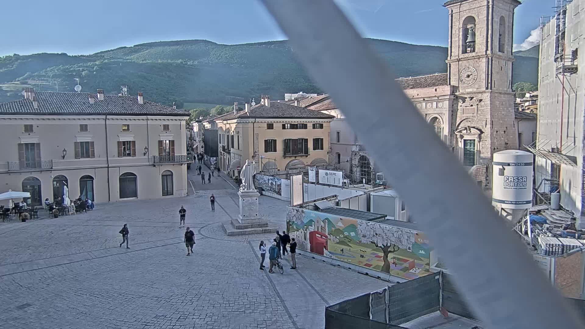 A town square with several people walking around, buildings surrounding it, a church with a bell tower in the background, and mountains under a clear sky.