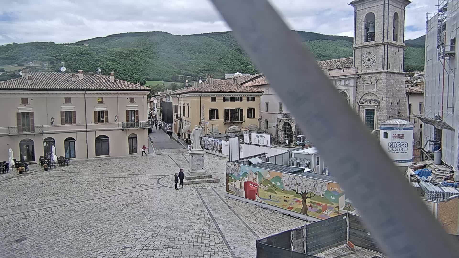 A cobblestone town square, partially obscured by a construction site, sits beneath a cloudy sky, with mountains in the background.