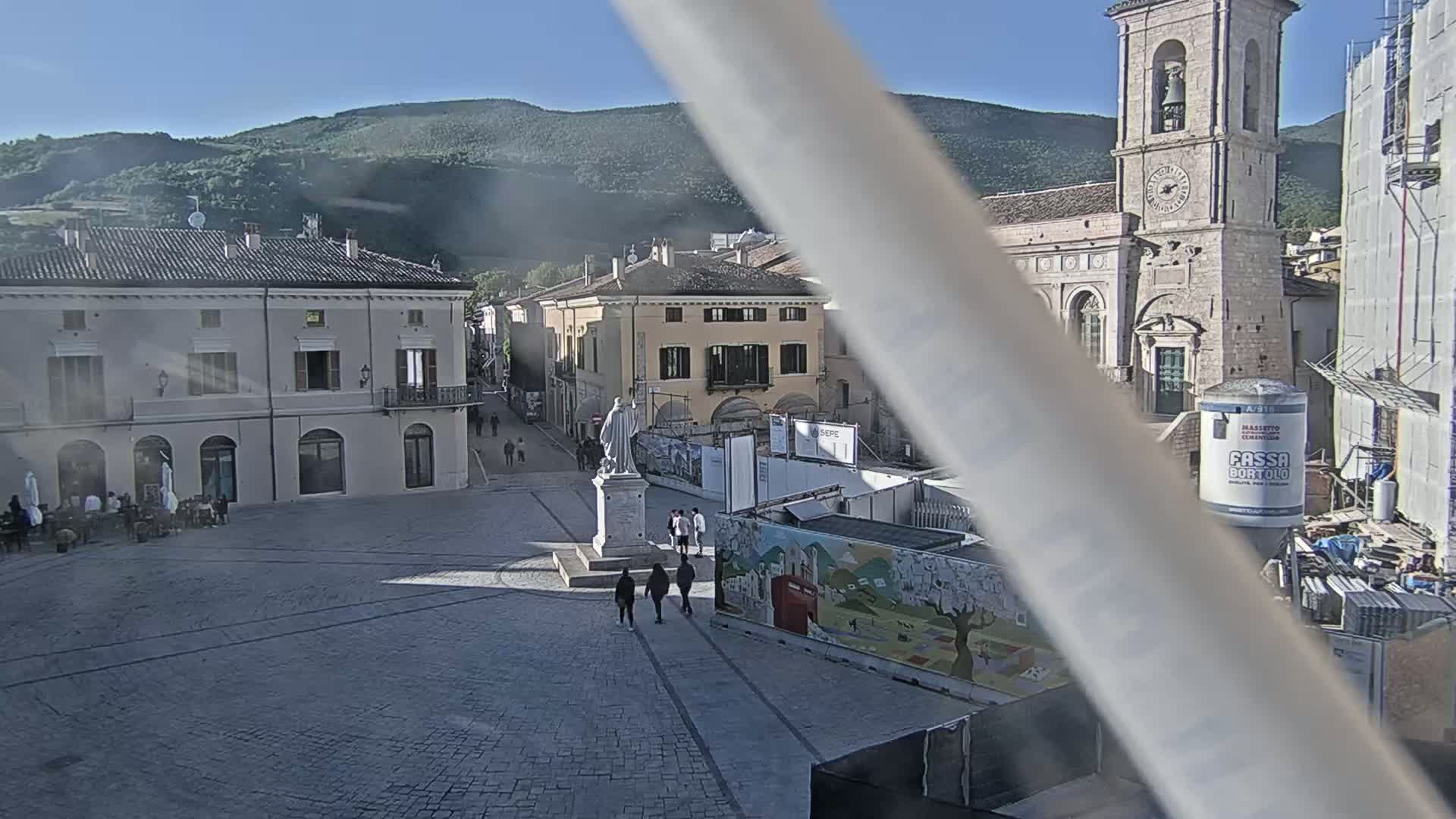A sunny day reveals a town square with several buildings, a statue, and some construction, all set against a backdrop of mountains.