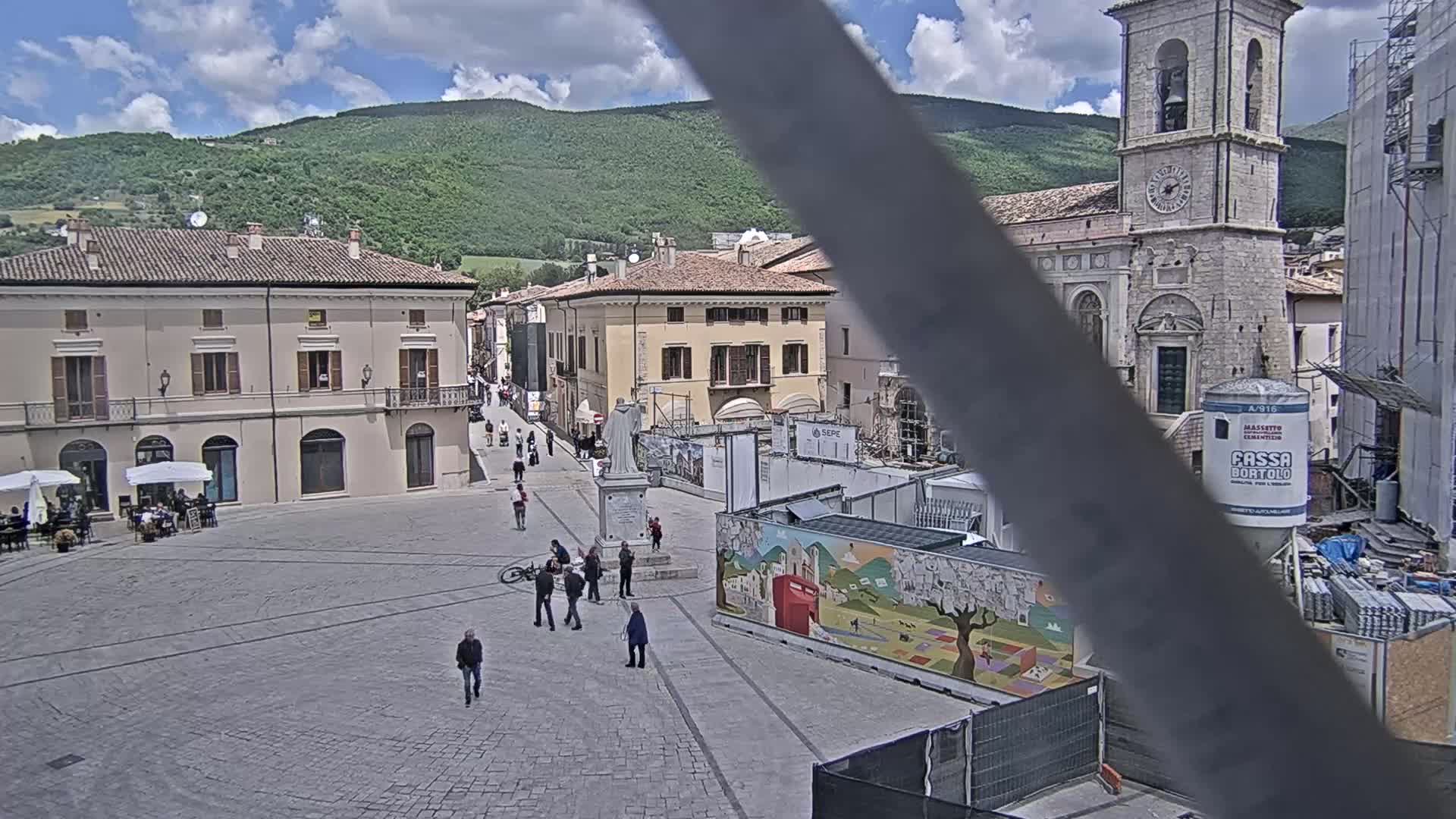 A town square with several buildings, a statue, some people, and construction, under a partly sunny sky.