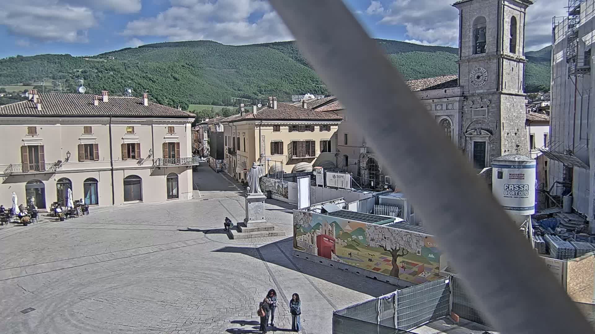 A mostly empty town square, under a partly cloudy sky, is surrounded by buildings, including a tall church, with construction occurring in a section of the square.
