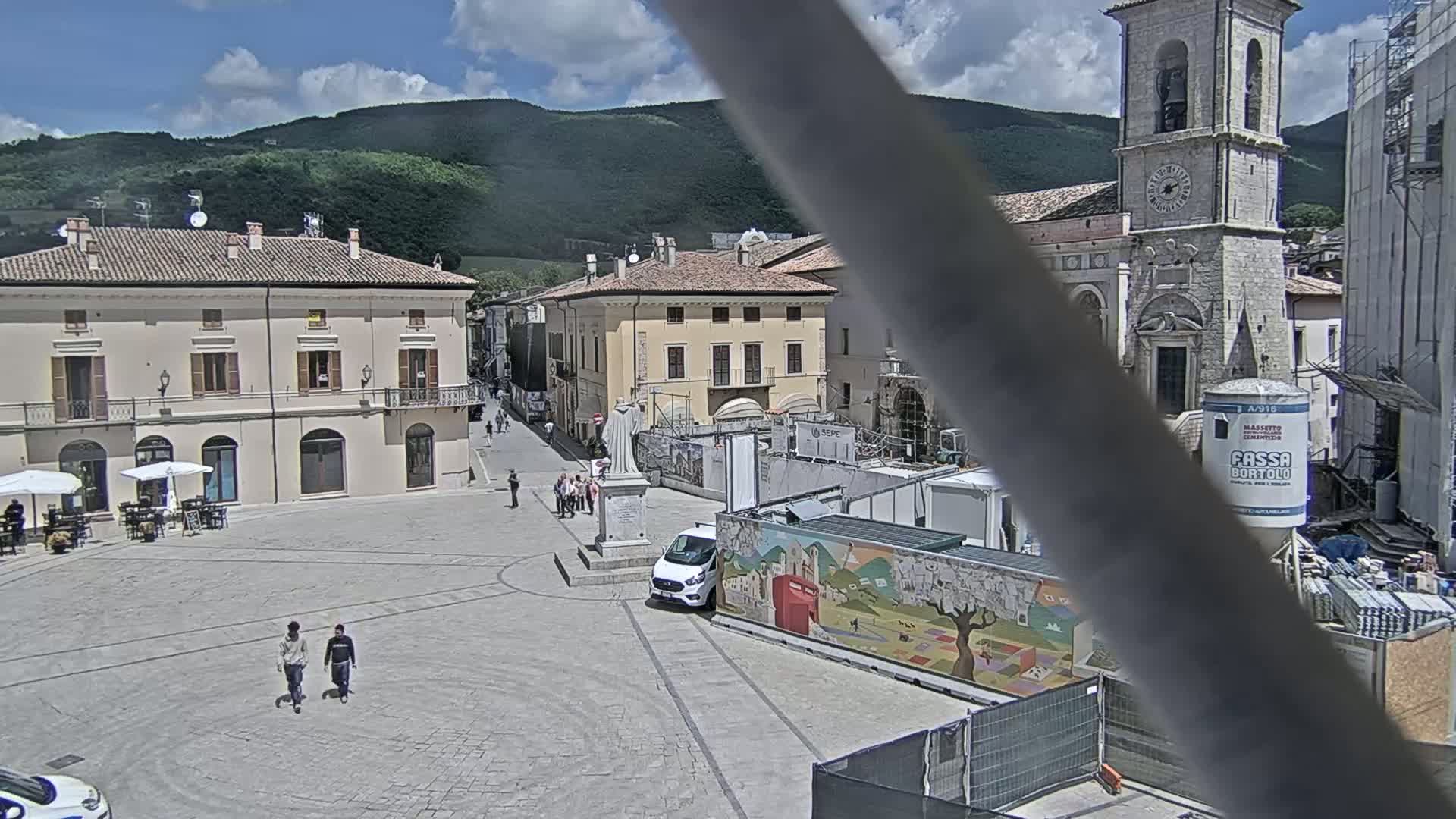 A town square with several buildings, including a church with a tall bell tower, is under construction on a sunny day with partly cloudy skies and green hills in the background.