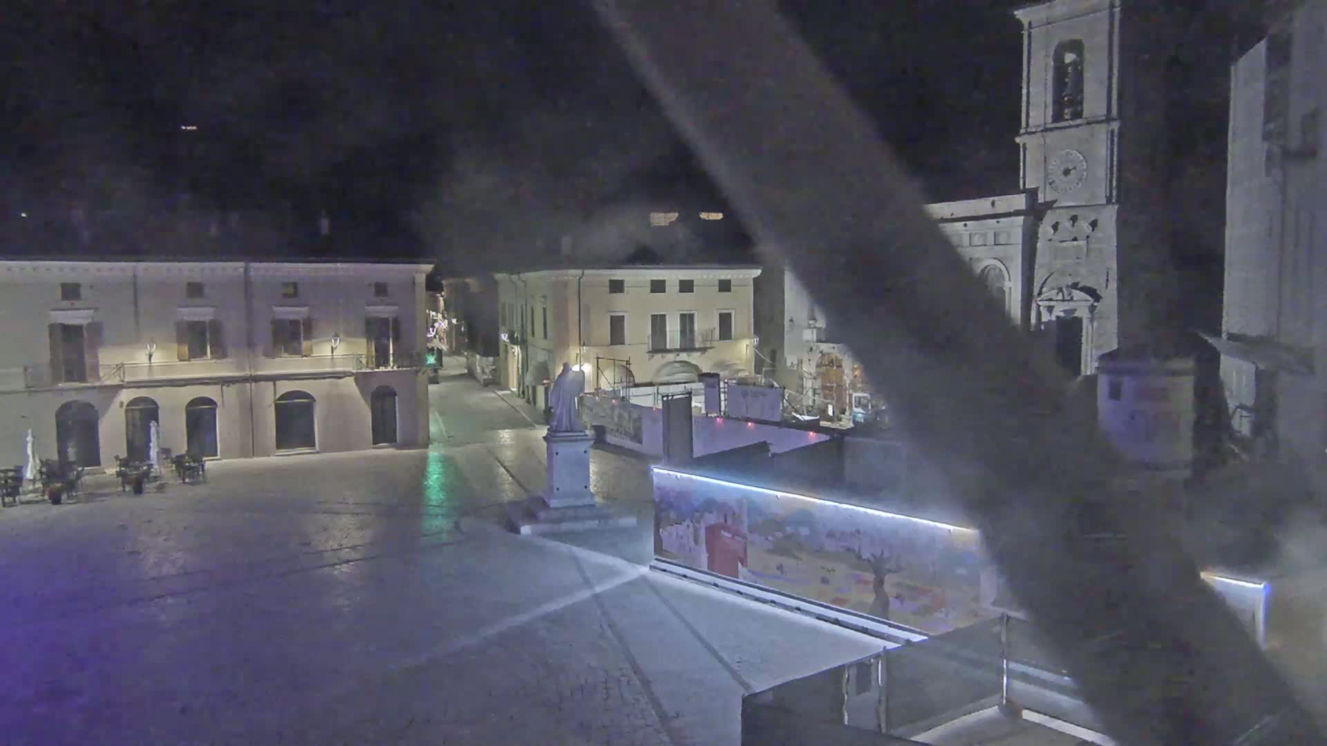 A nighttime, high-angle view of a town square with buildings, a statue, and construction barriers.