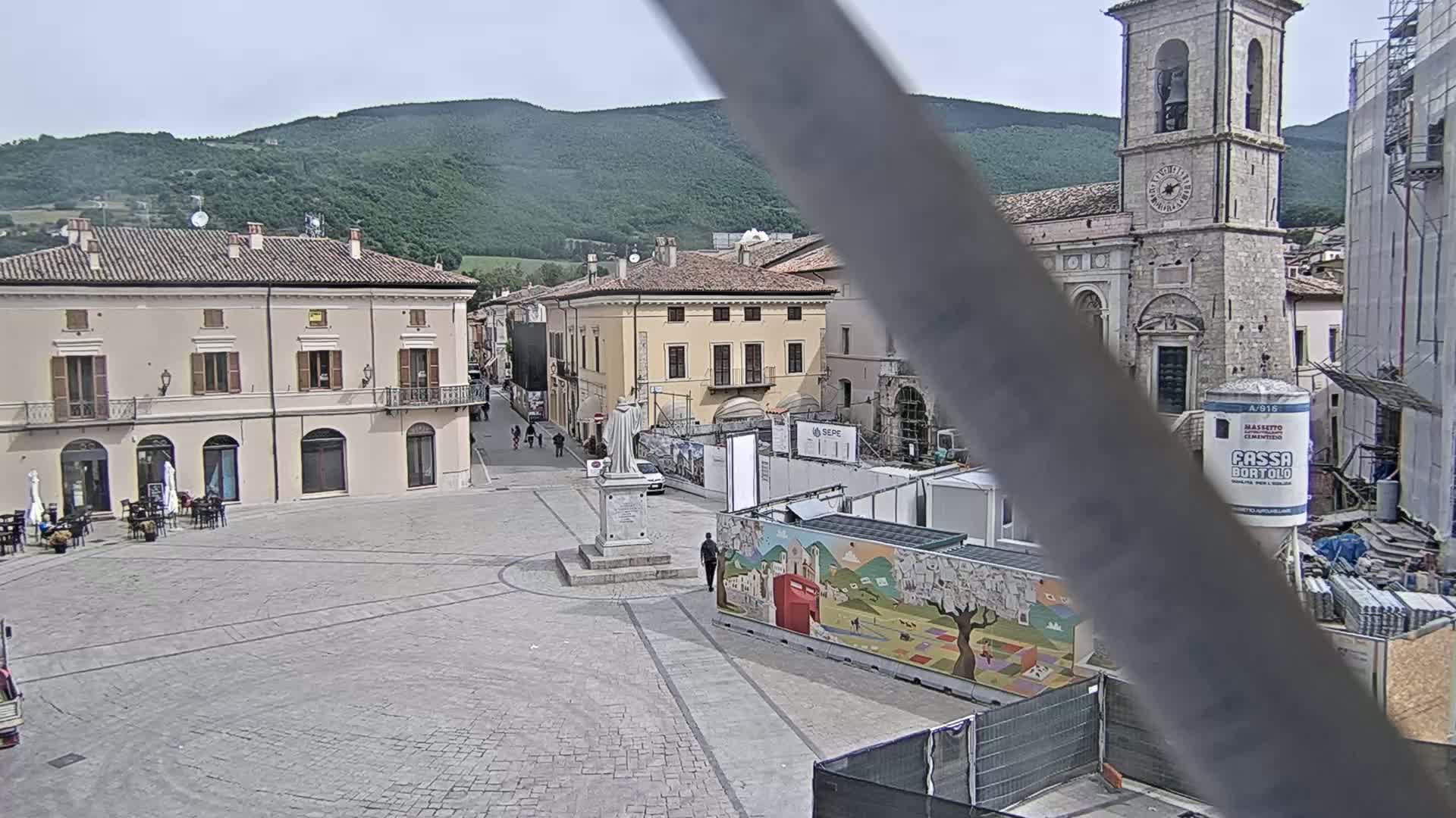 A town square with several buildings, a statue, and construction in progress, all under a partly cloudy sky with mountains in the background.