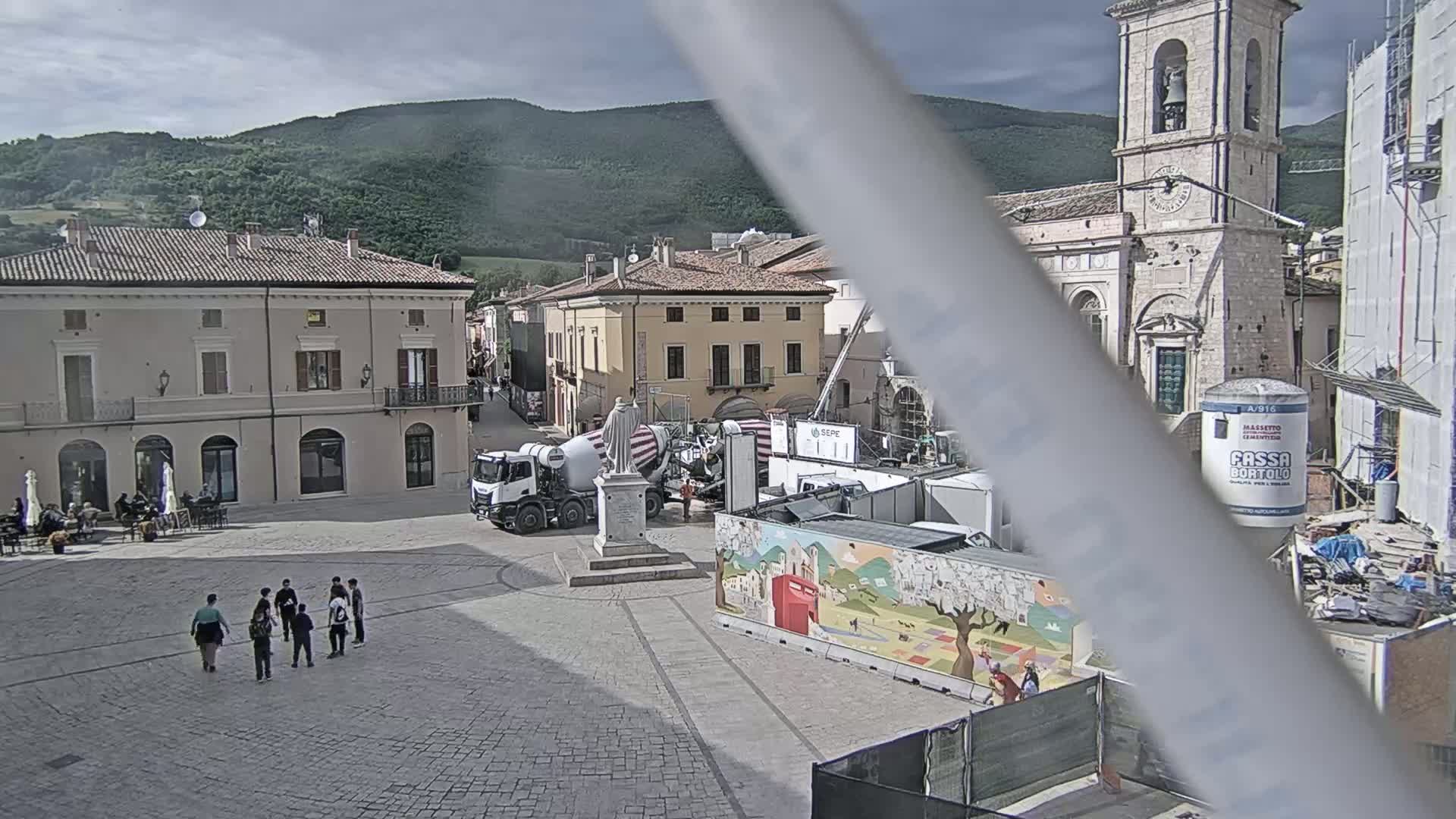 A town square under partly cloudy skies is partially obscured by construction, including a cement mixer and a painted barrier, with several people standing near a statue in the foreground.