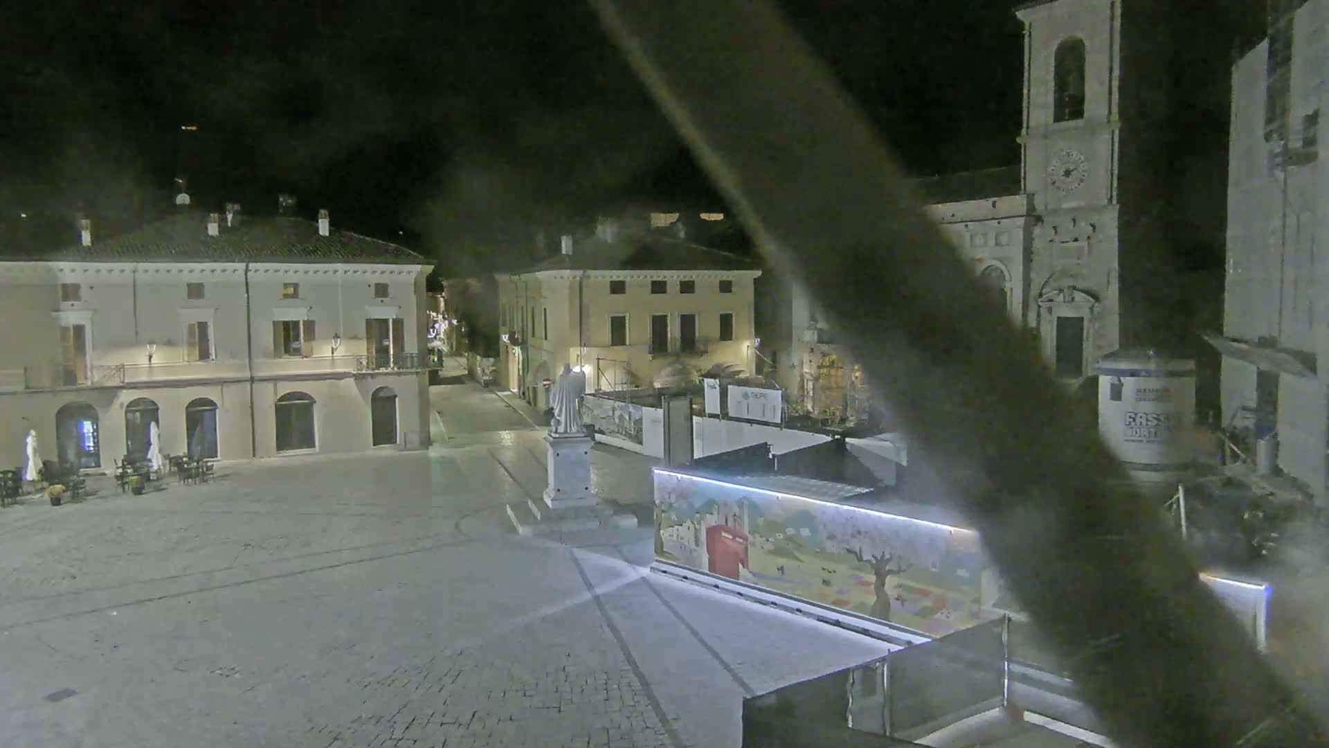 A nighttime view of a town square with several buildings, a statue, and construction barriers surrounding a brightly lit mural.