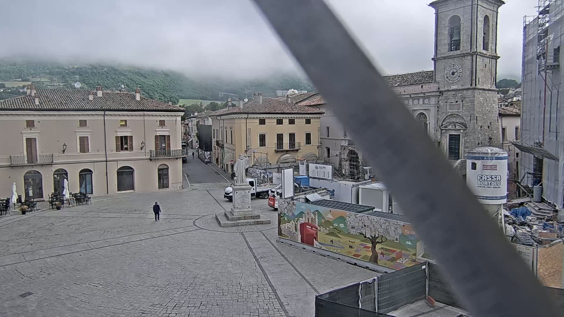 A stone plaza with several buildings, a statue, construction equipment, and a person walking, all under an overcast sky with mountains in the background.