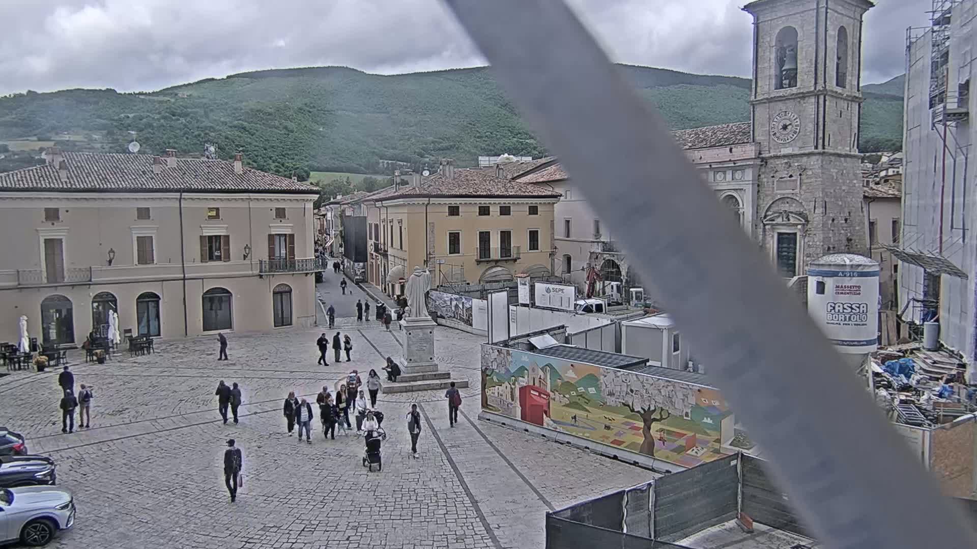 A town square with several people walking around, buildings surrounding it, construction in progress, and a partially cloudy sky over green hills in the background.