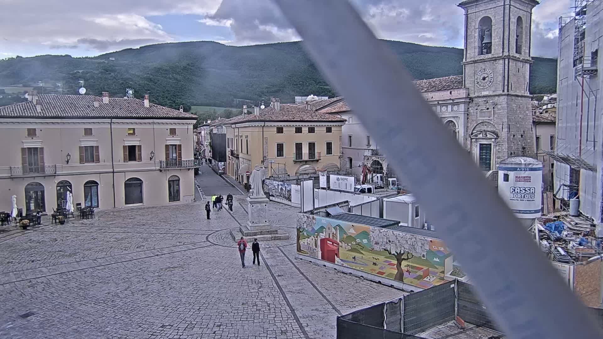 A cobblestone town square is partially obscured by a construction site and features several buildings, a statue, and a few pedestrians under overcast skies with mountains in the background.