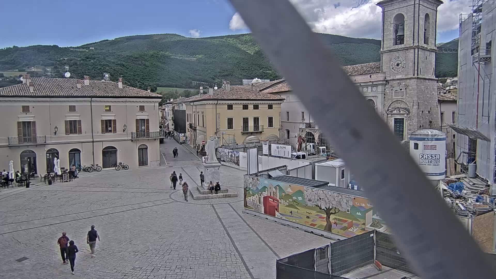 A mostly empty town square with several buildings, including a church tower, under a partly cloudy sky, with construction underway in parts of the square.