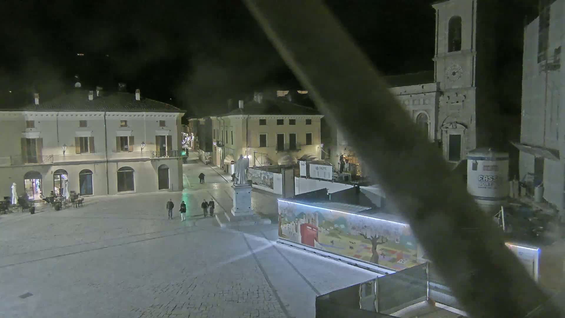 A nighttime view of a town square shows several buildings, a statue, some pedestrians, and construction materials, under dark conditions.
