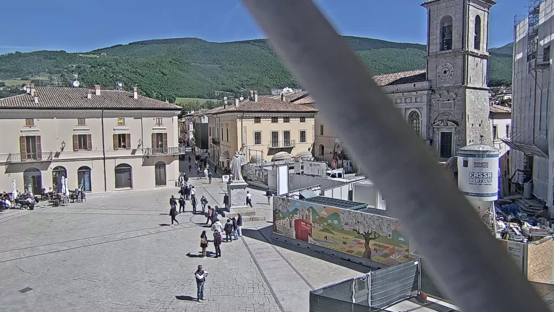 A town square with several buildings, a church, and people walking around on a sunny day with mountains in the background.
